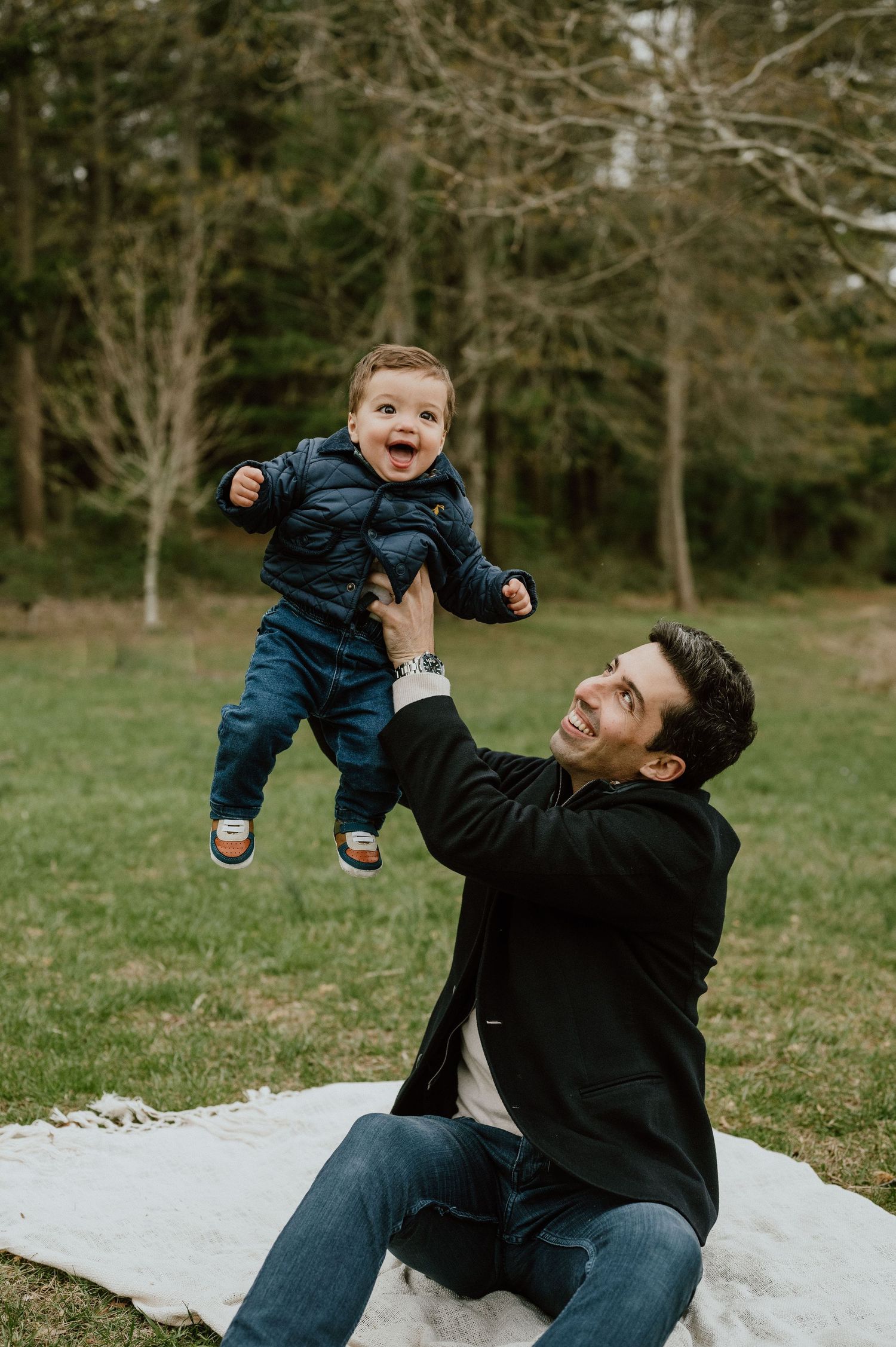 Parent lifts laughing toddler in navy jacket during outdoor photoshoot.