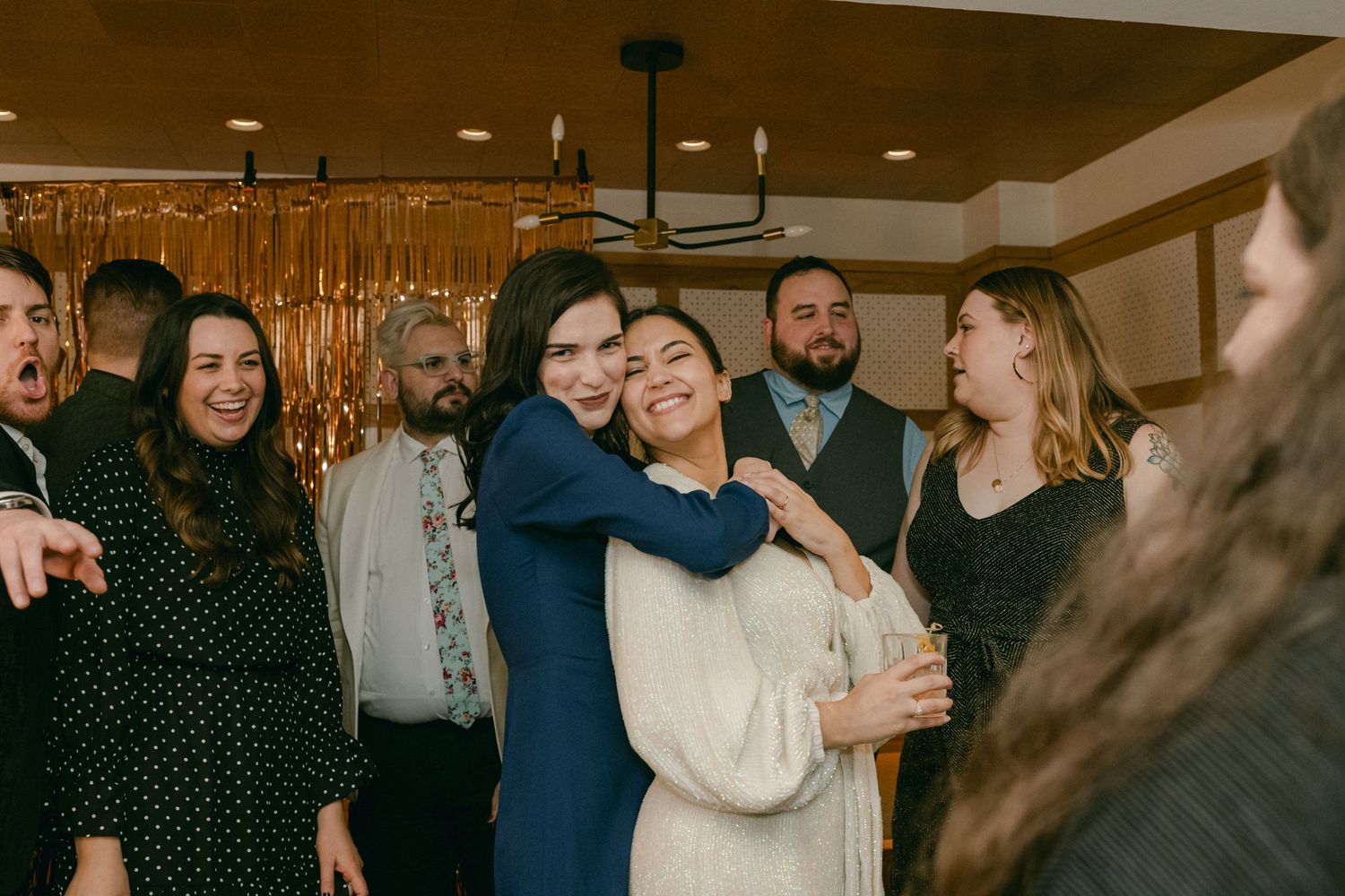 Wedding celebration with guests gathered around embracing couple at indoor reception venue.