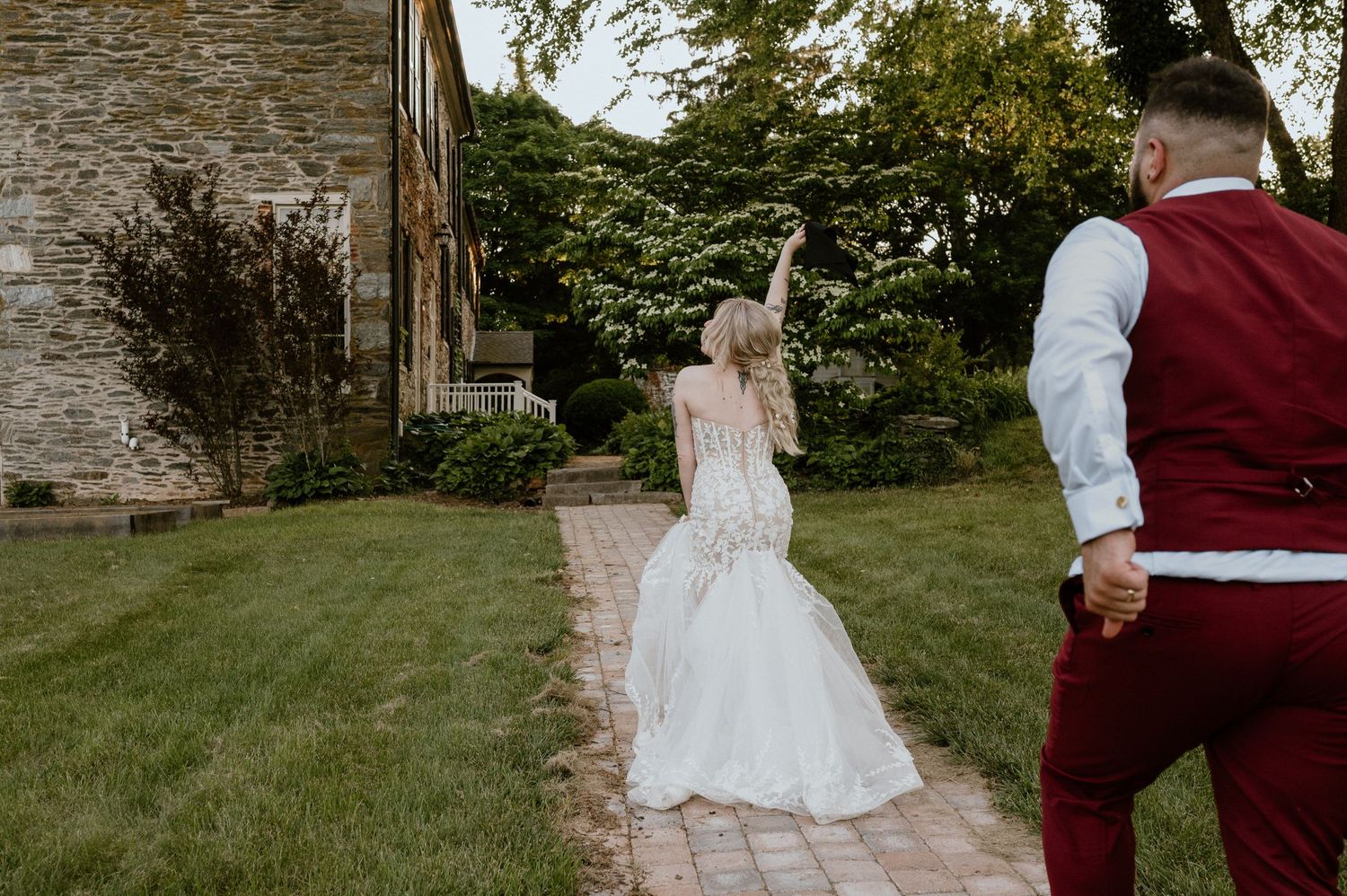 Bride in lace gown poses playfully on garden path with groom in burgundy vest.