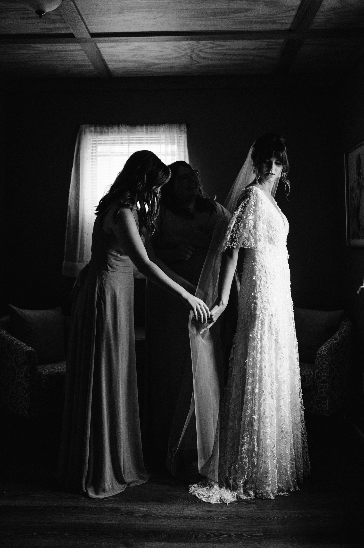Silhouetted figures help adjust a wedding dress in dramatic window lighting.