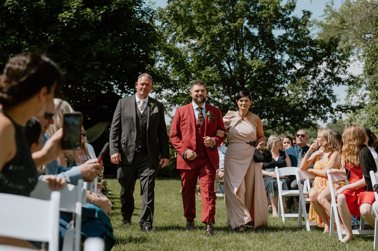 Wedding party walking down aisle at outdoor ceremony with guests seated on white chairs.