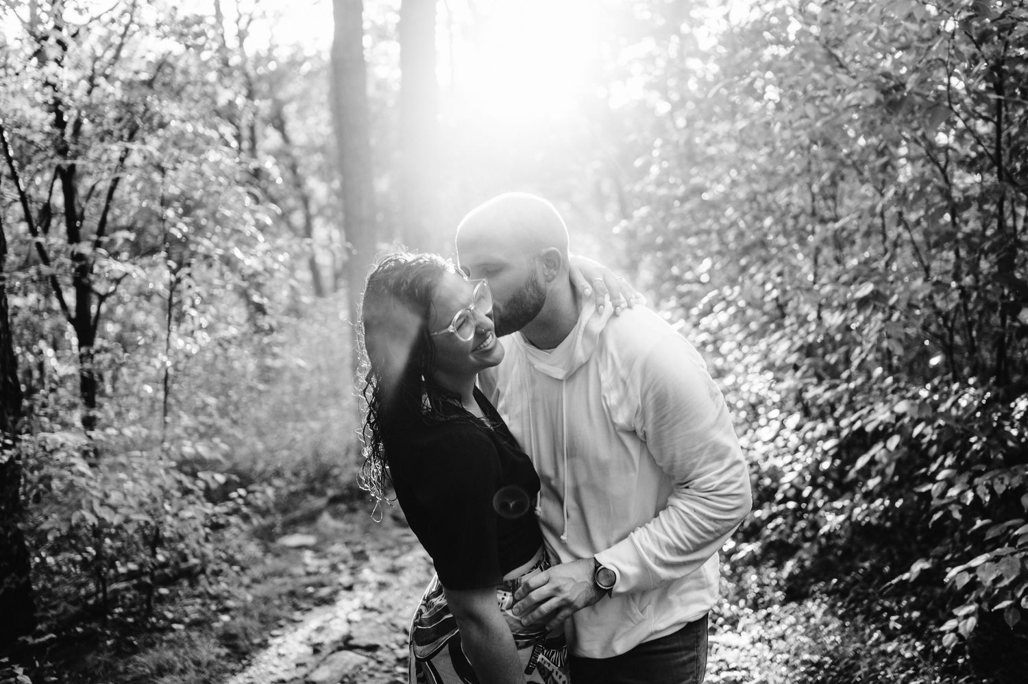 Black and white photo of couple sharing tender moment on wooded trail.