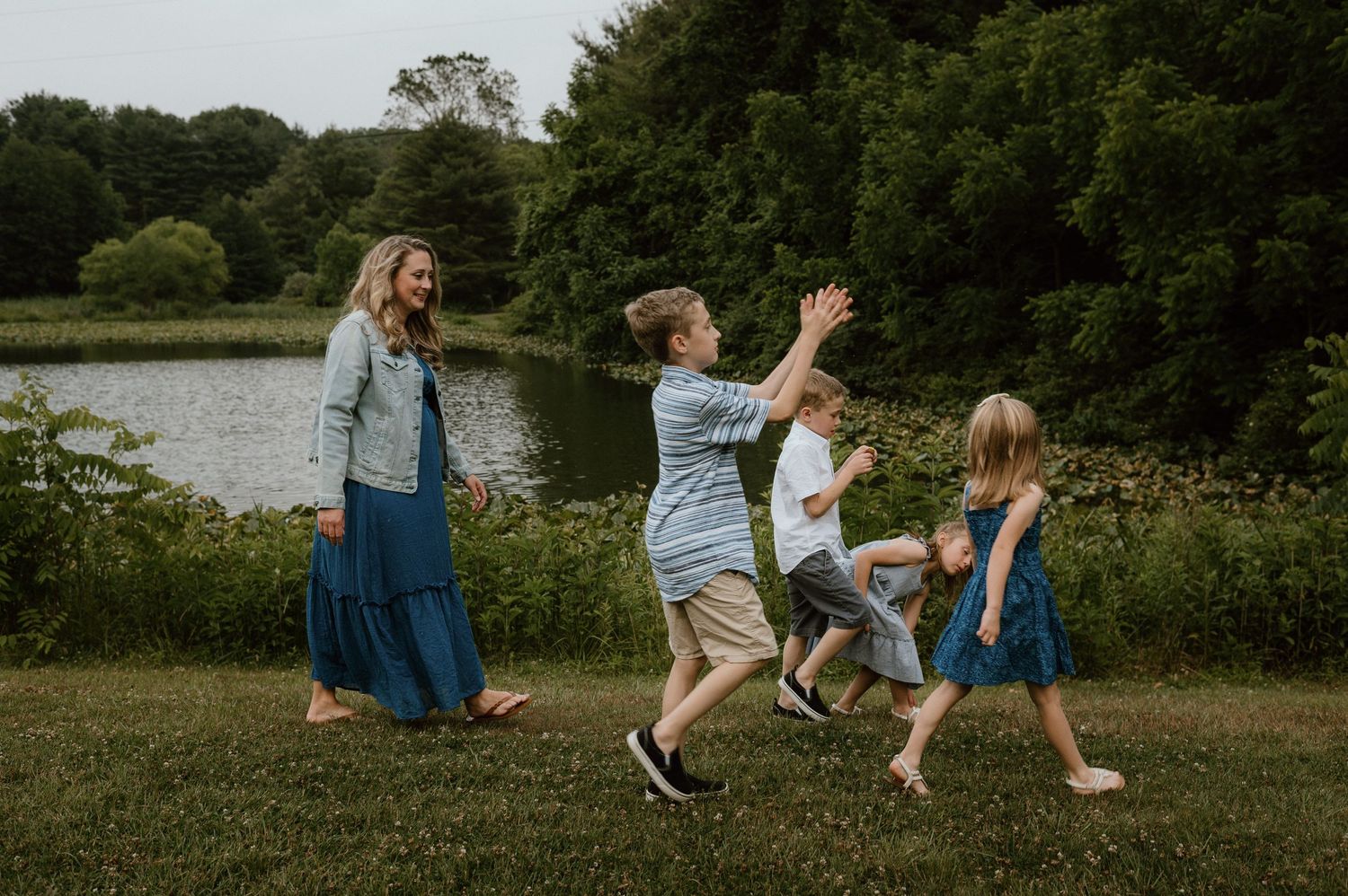 A group dances and plays together on grass near a lake and trees on a summer day.