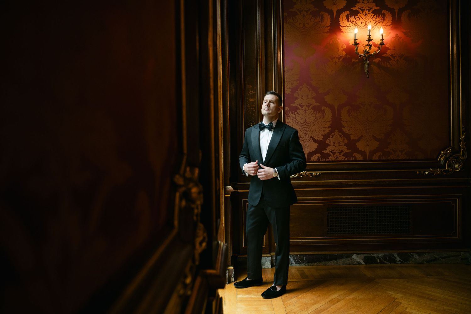 An elegant person in a black tuxedo stands in a luxurious room with ornate walls and warm lighting from a wall sconce.