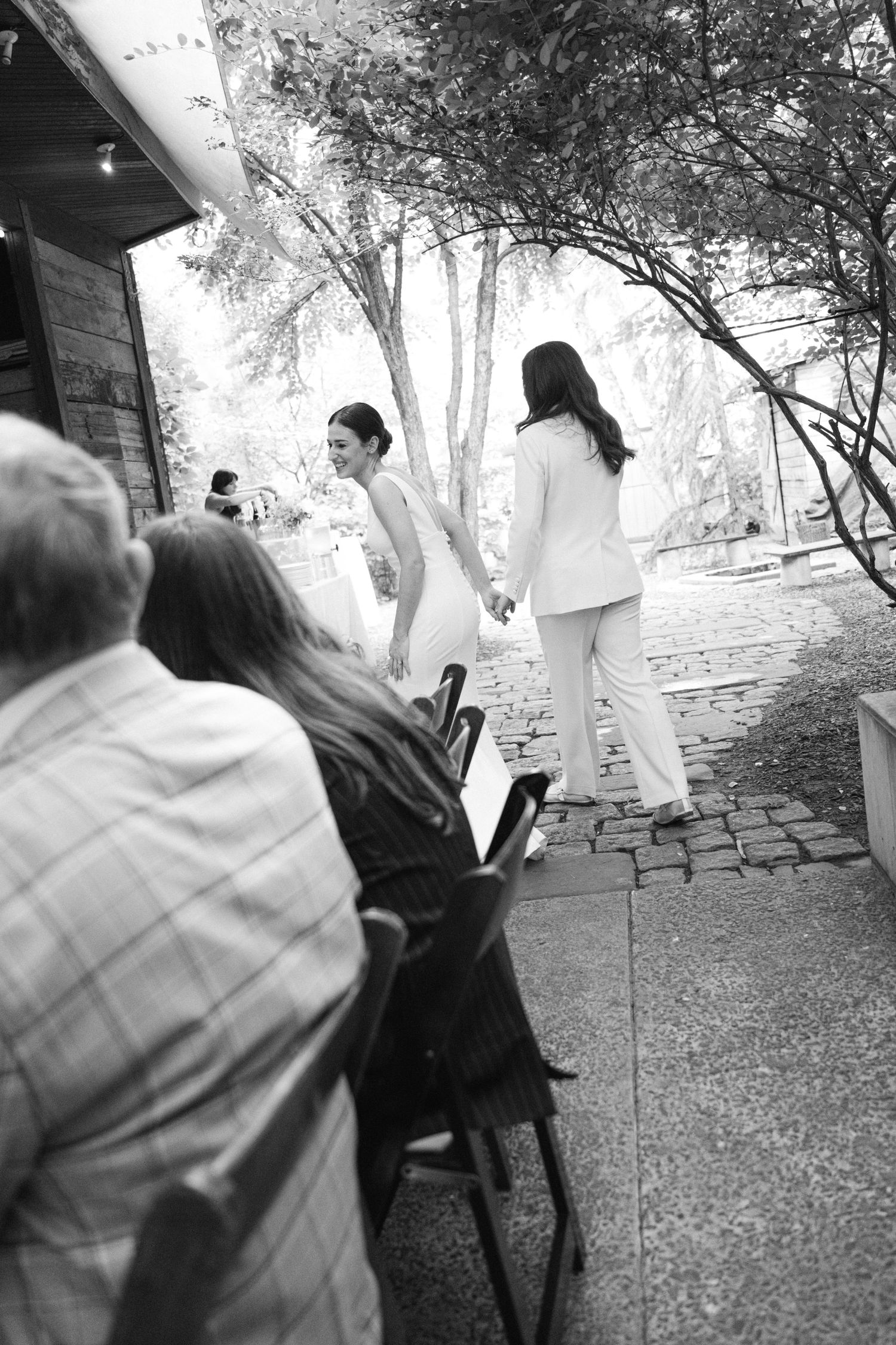 Black and white photo showing guests at an outdoor wedding ceremony on a stone path under trees.