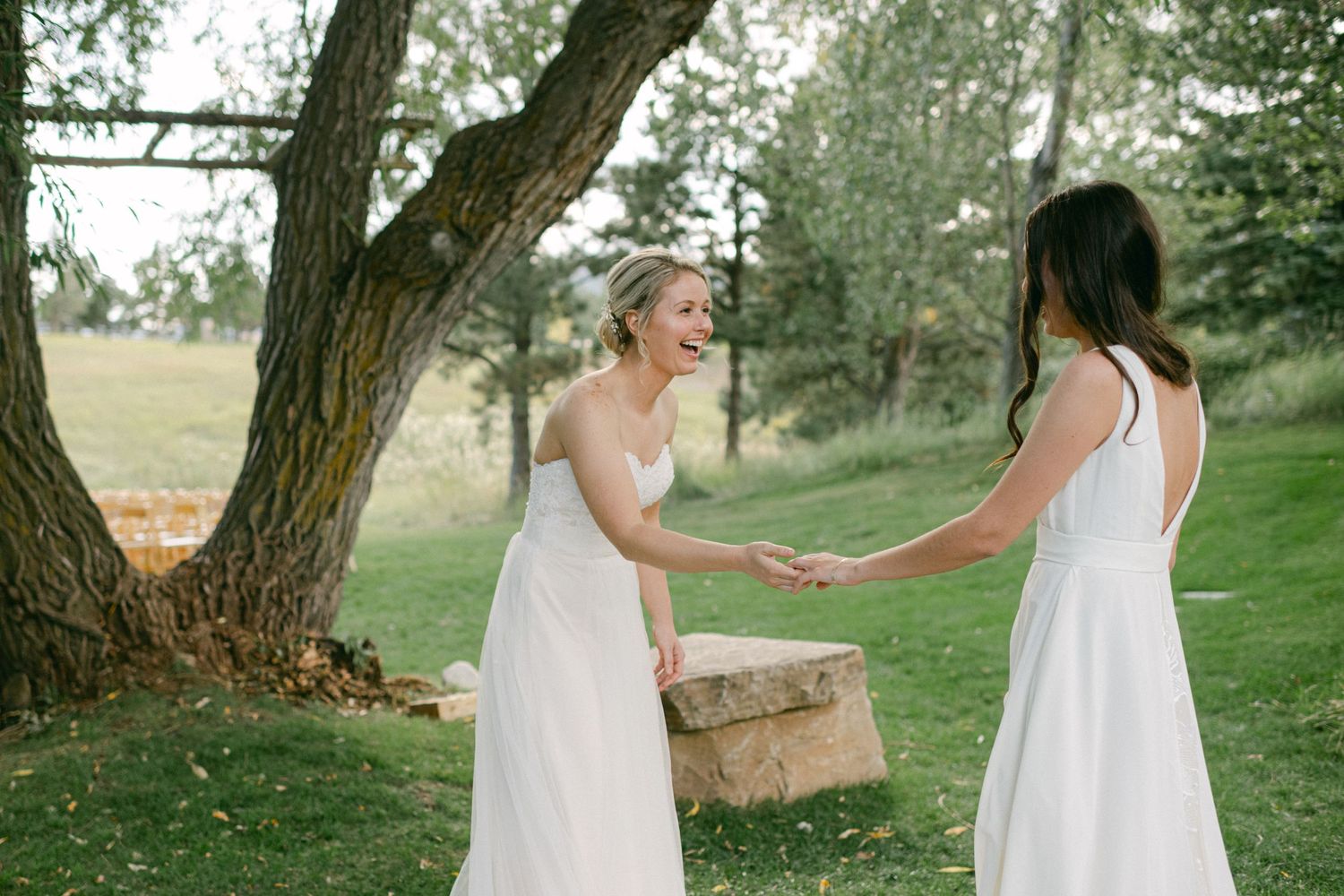Two women in white dresses laughing together under tree in outdoor setting.