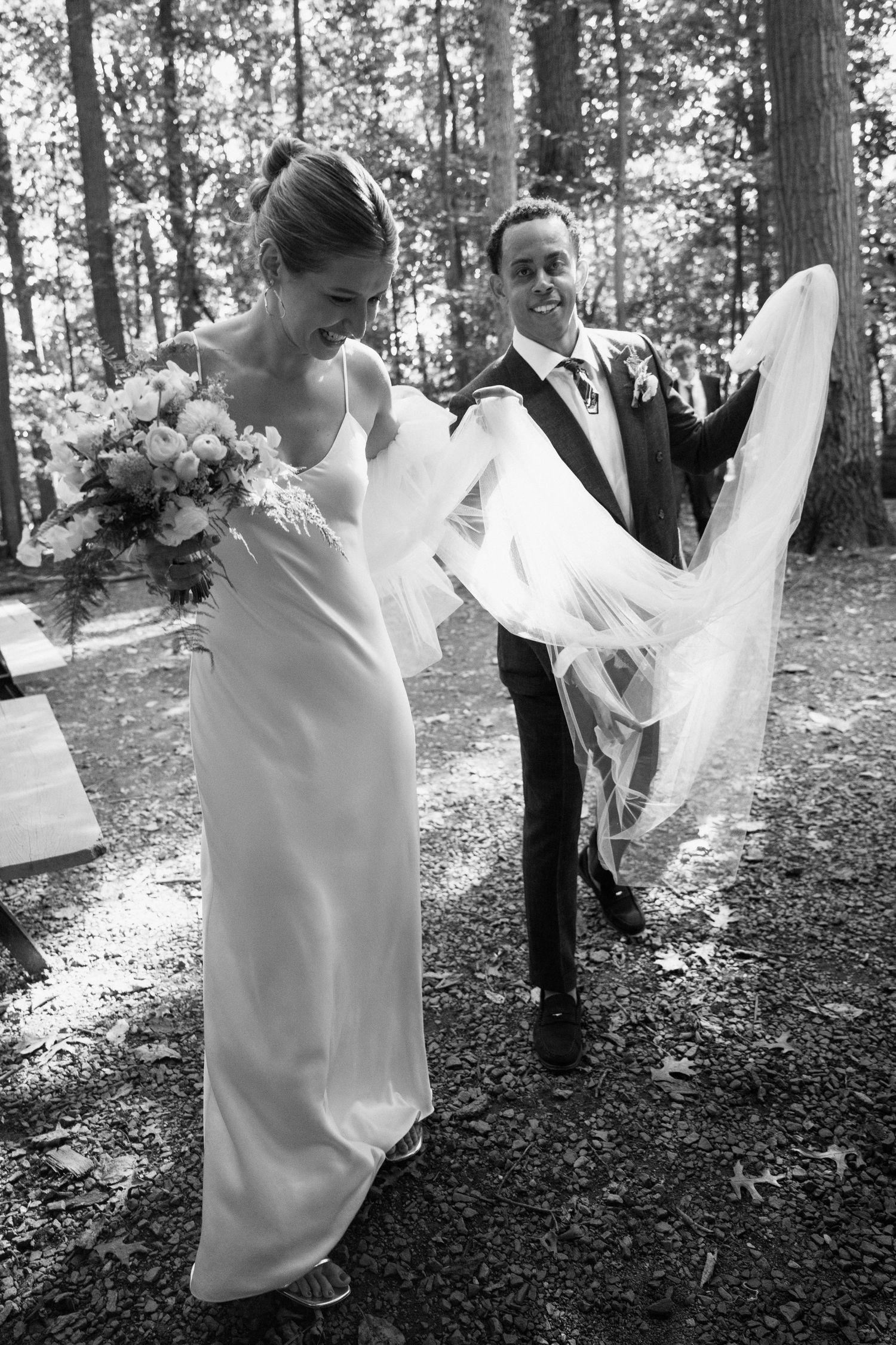 The couple walks together through a forest setting carrying a bouquet of white flowers.