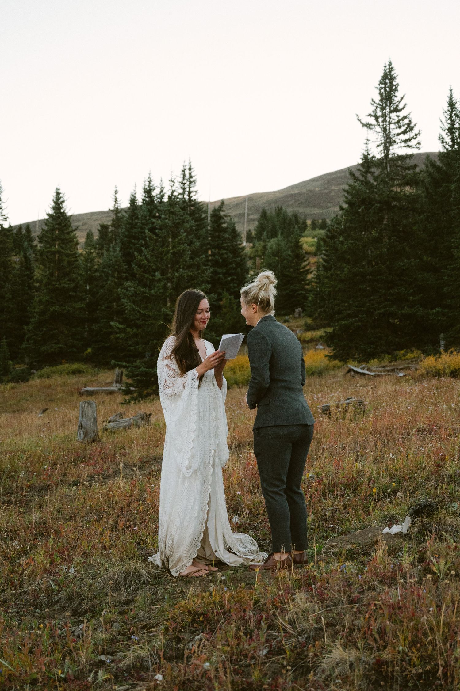Two people exchange vows in mountain meadow surrounded by pine trees at sunset.