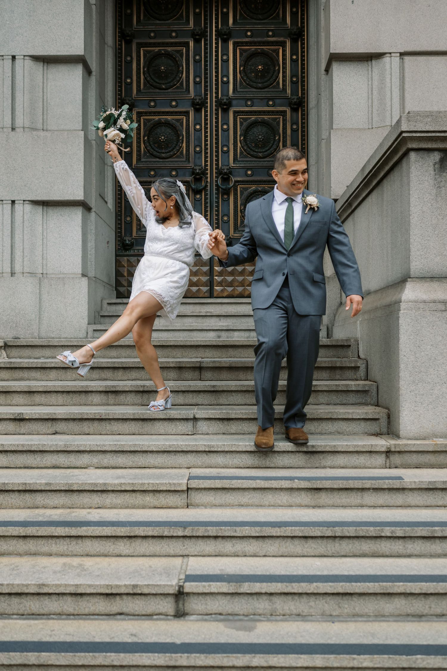 A joyful couple celebrates on stone steps outside an ornate building with decorative doors.