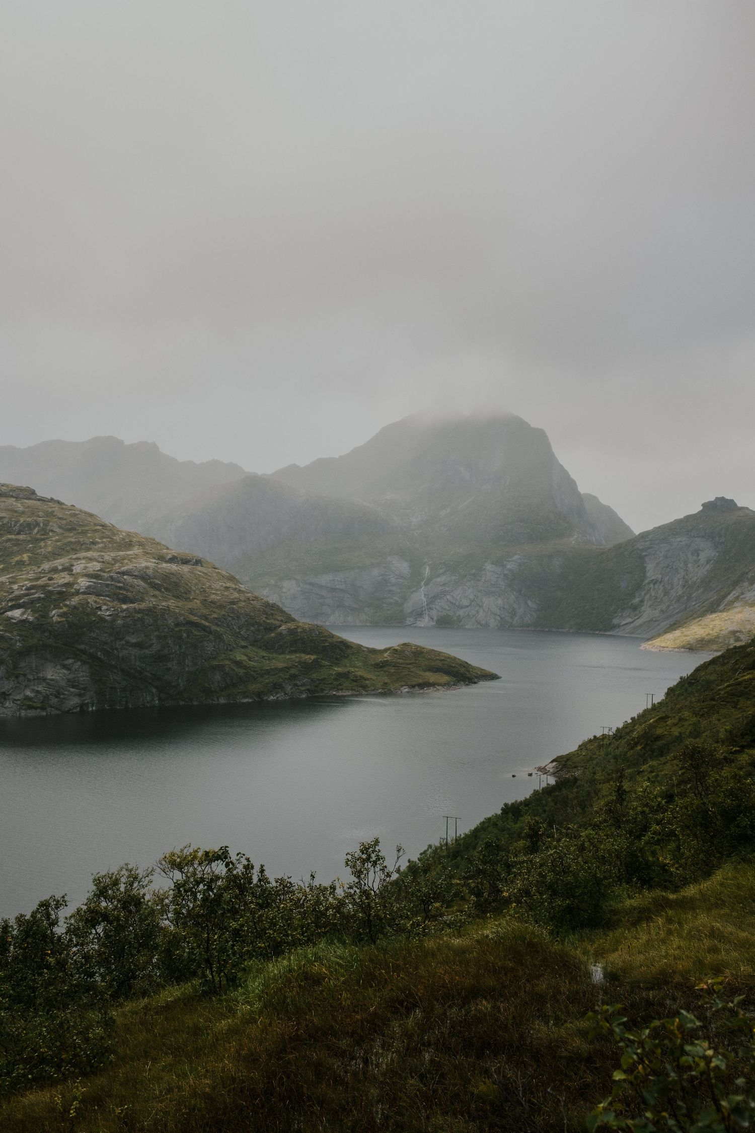 Misty mountain lake surrounded by dramatic peaks and lush green slopes in moody weather.
