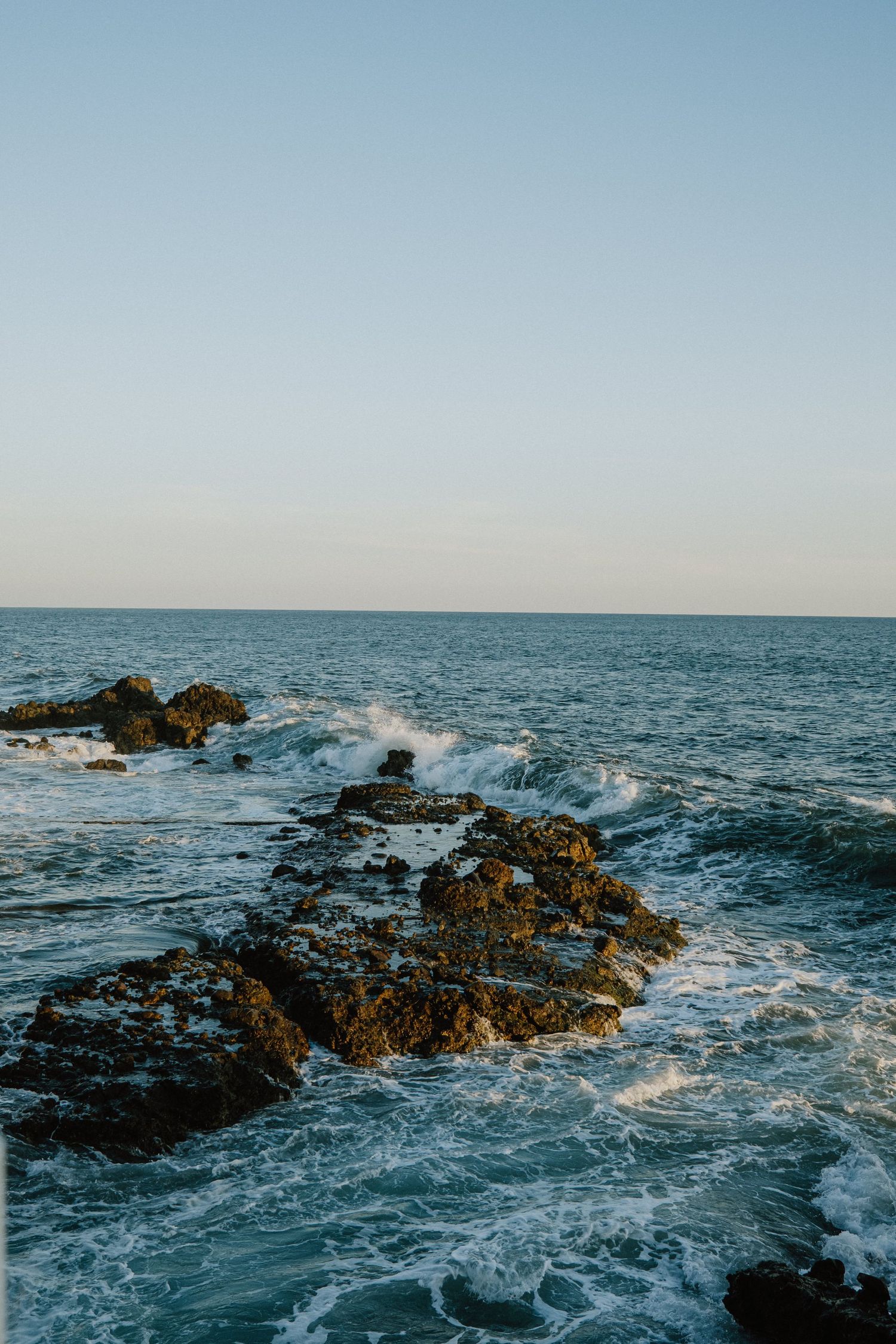 Waves crash against rocky outcrops in the blue ocean under clear skies.