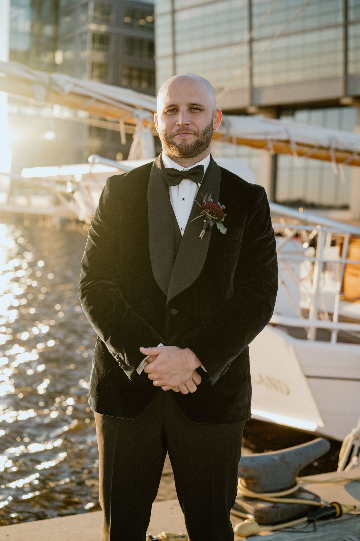 Groom in classic black tuxedo poses at waterfront venue during golden hour.