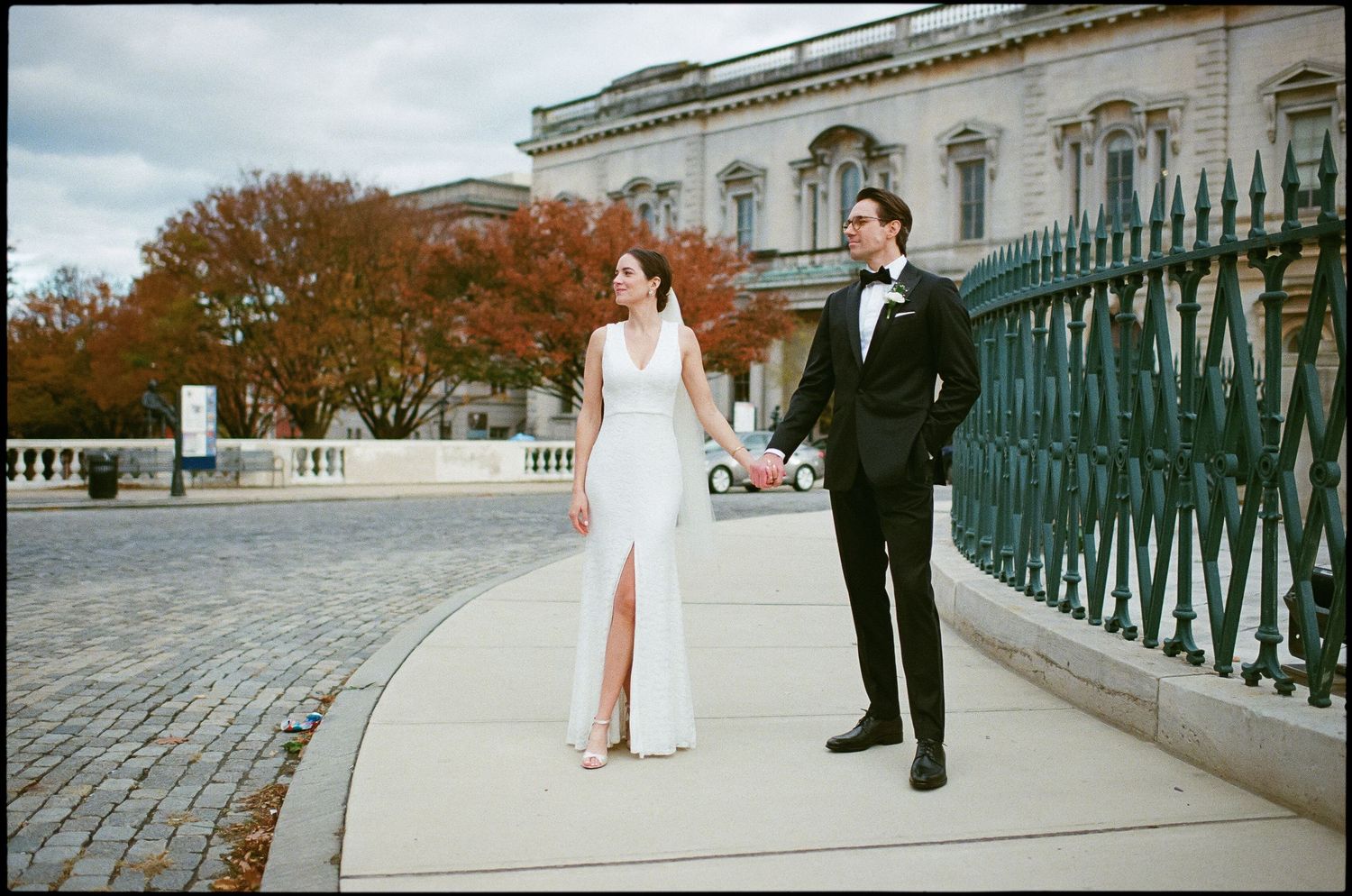 Newlyweds walking along an ornate railing against autumn trees and classical architecture background.