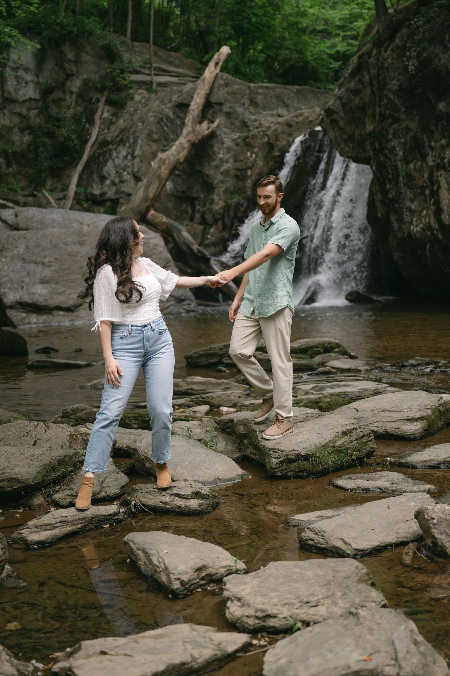 Two people carefully navigate across rocky stepping stones near a forest waterfall.