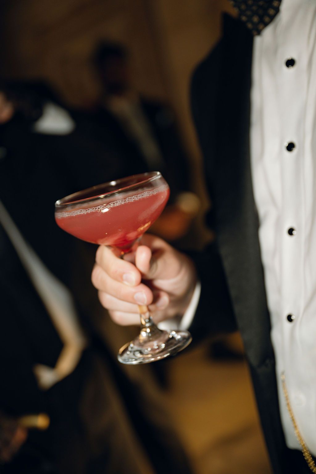 Several hands hold pink cocktails in coupe glasses against a dark background at what appears to be an elegant event.