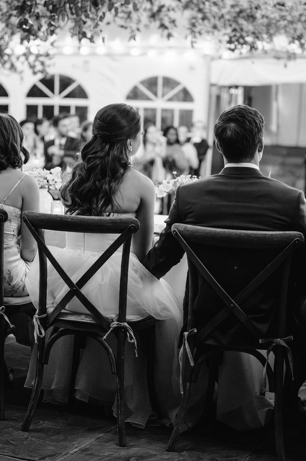 Row of guests seated in cross-back chairs at an outdoor wedding reception.