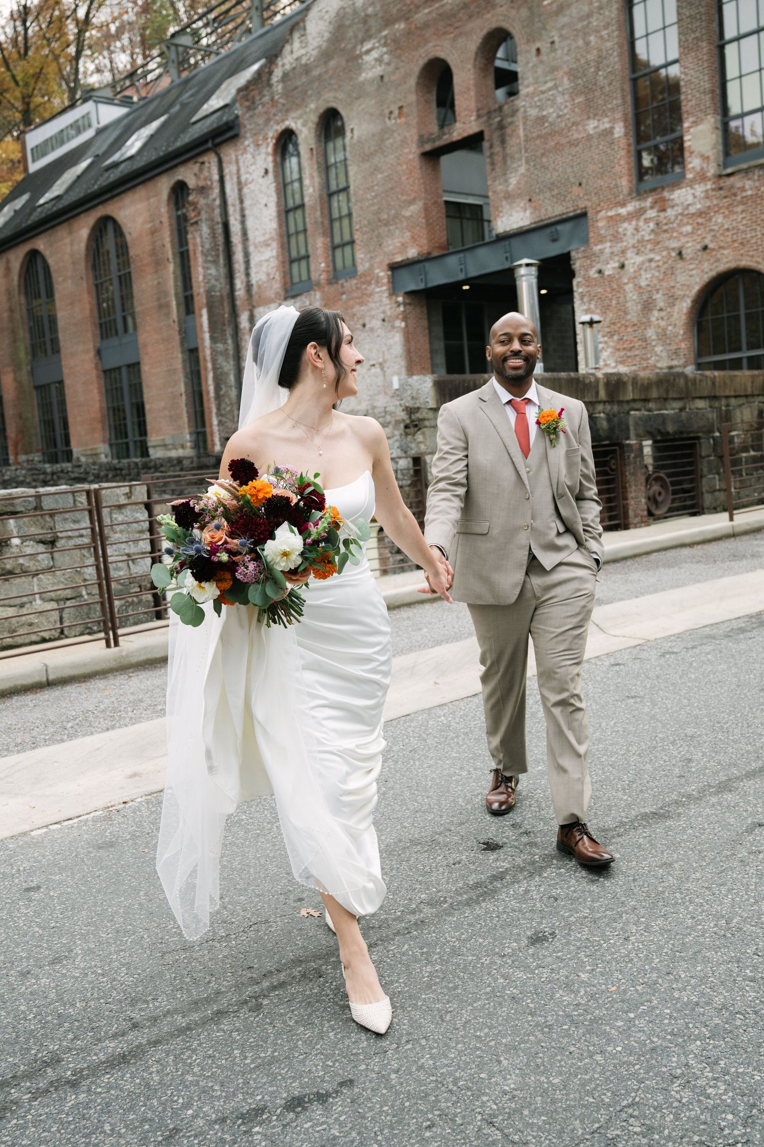 A bride in a white gown holding a colorful bouquet walks hand-in-hand with a groom in a tan suit on a city street.