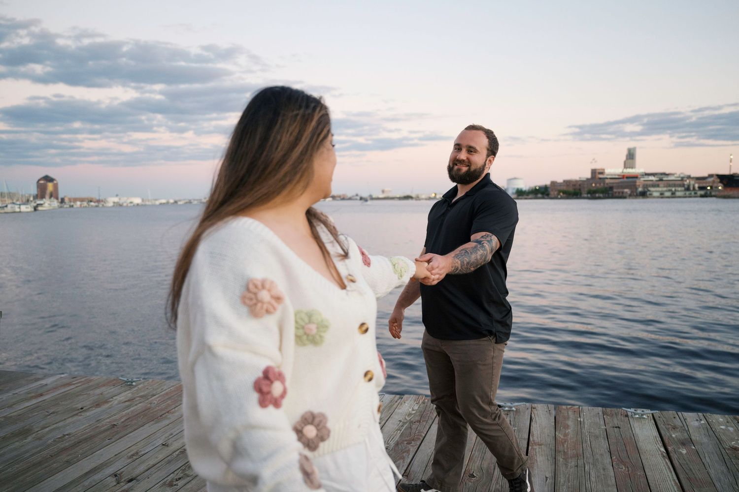 A couple shares a romantic moment on a wooden pier overlooking a waterfront cityscape at dusk.