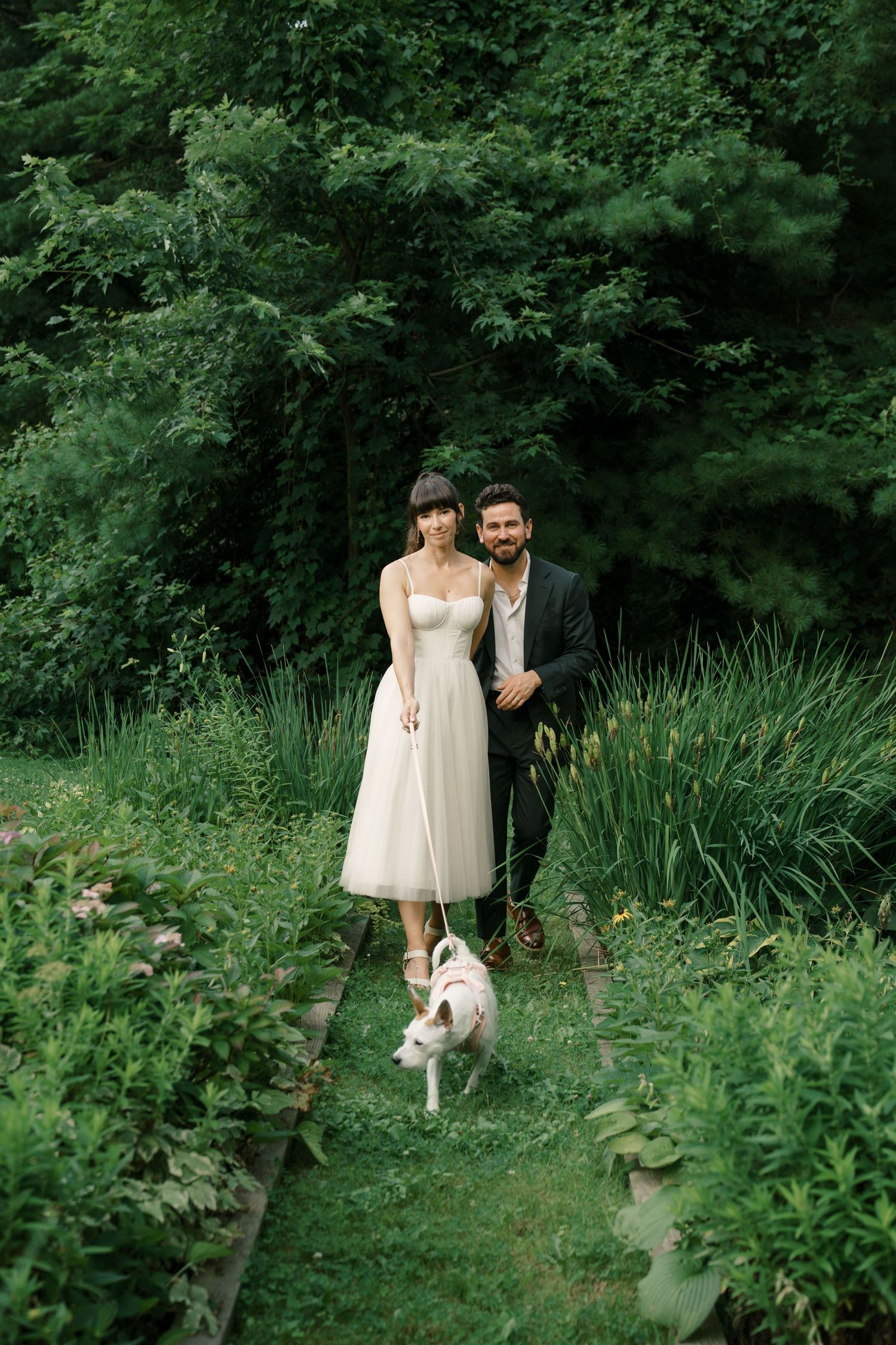 Couple standing on garden path with their dog among lush greenery and irises.