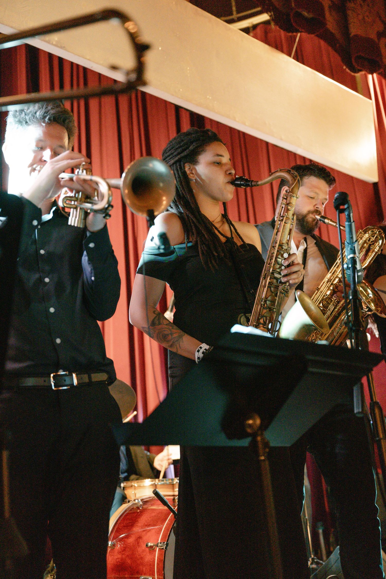 Musicians performing live with trumpet, saxophone and vocals on stage with red curtain backdrop.