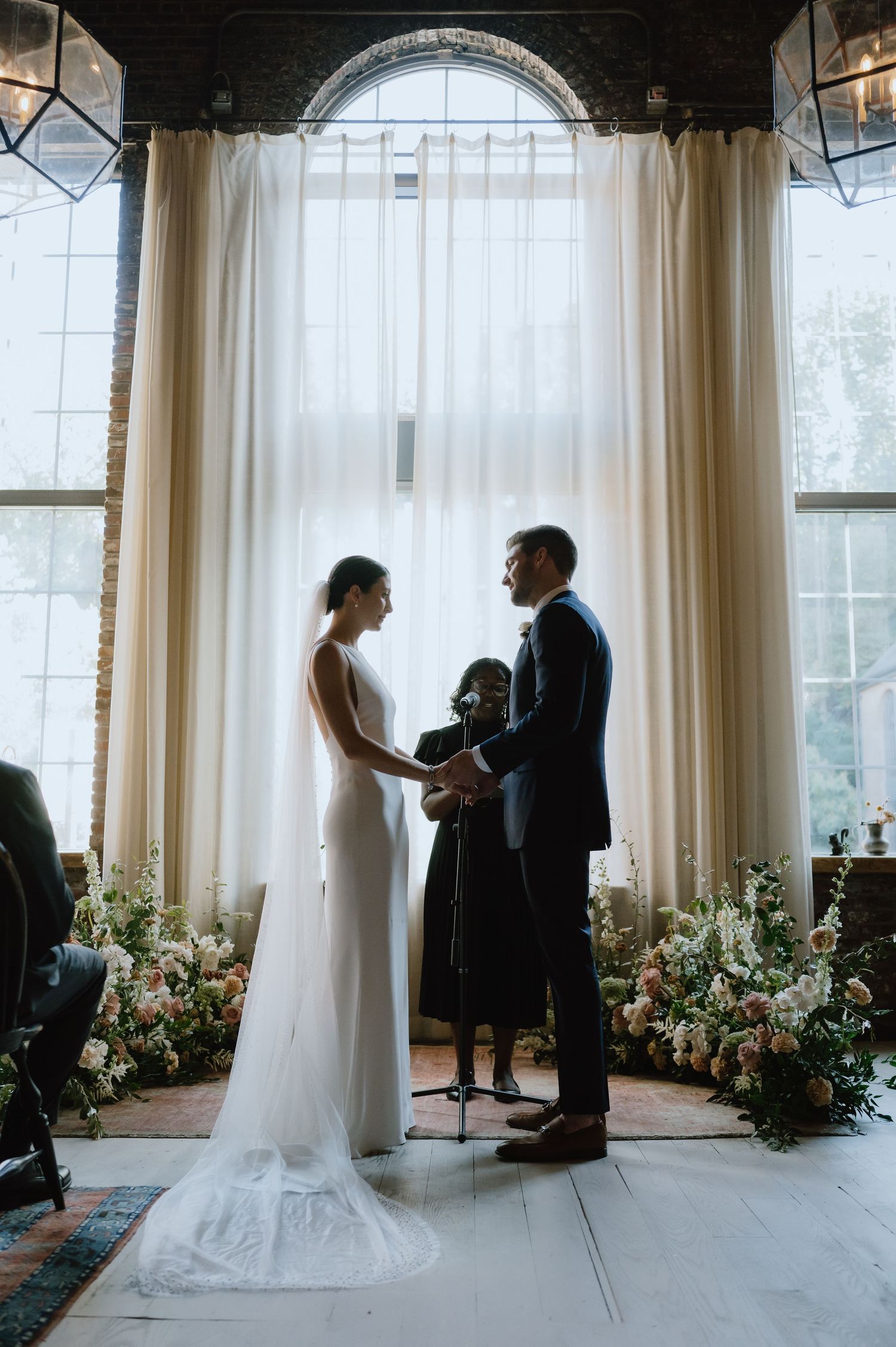 Wedding ceremony sequence in front of large windows with floral arrangements and sheer curtains.