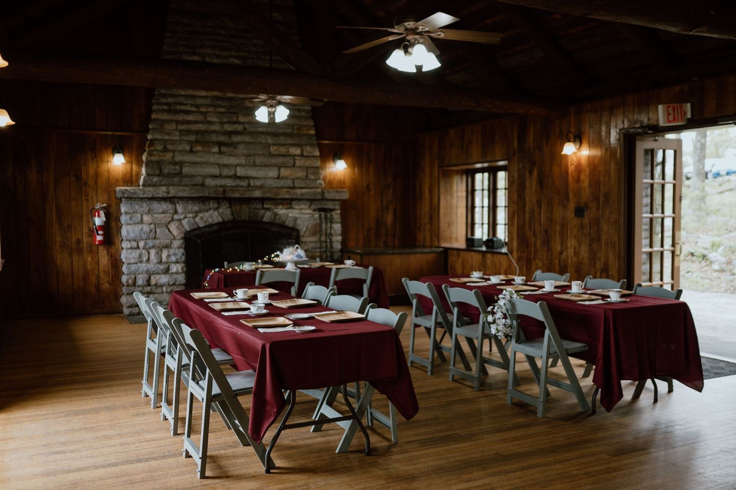 Rustic dining room with burgundy tablecloths and stone fireplace set for an elegant gathering.