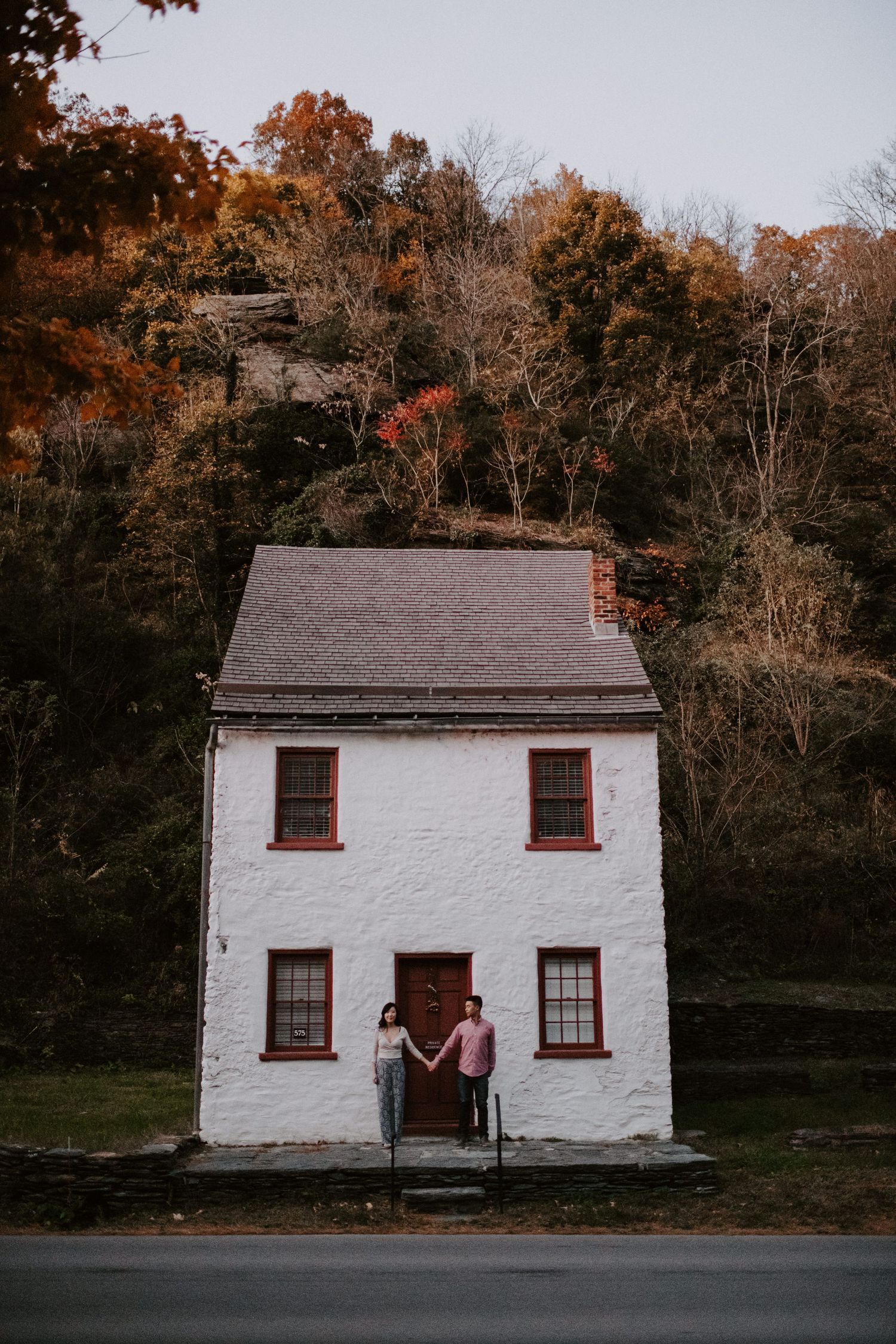 A couple sits in the doorway of a small white historic house during autumn.