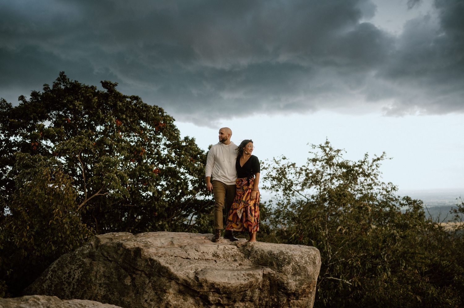 A couple poses together on a mountain outcrop with dramatic stormy skies behind them.