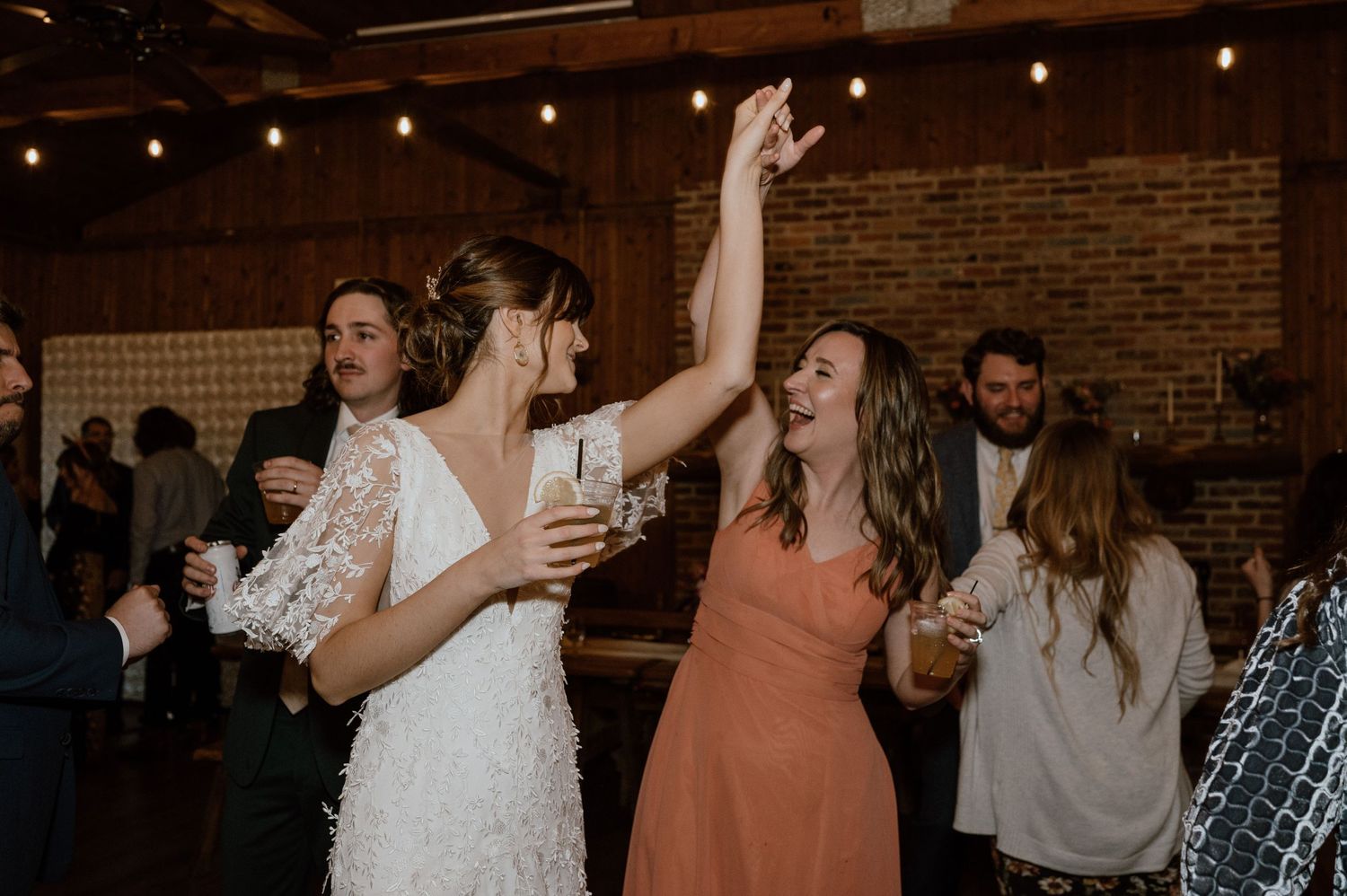 Wedding guests dancing and celebrating at a lively reception with brick walls and string lights.