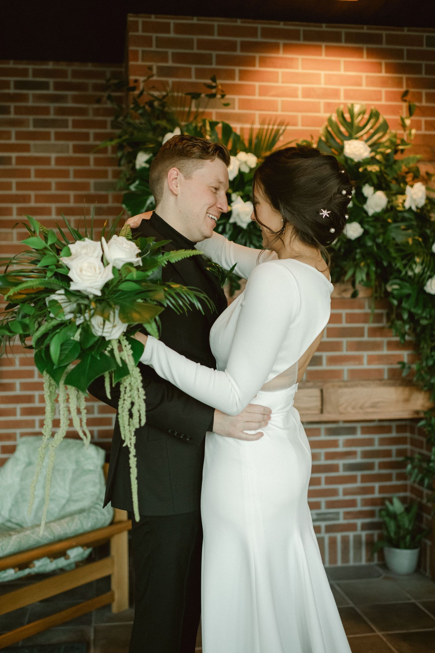 Romantic wedding portrait with couple embracing against brick wall and floral backdrop.