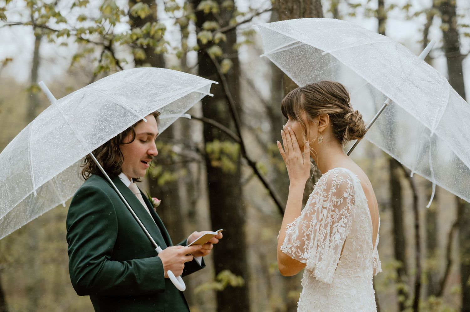 A couple exchanges vows while holding clear umbrellas in a forest setting during their rainy day wedding.