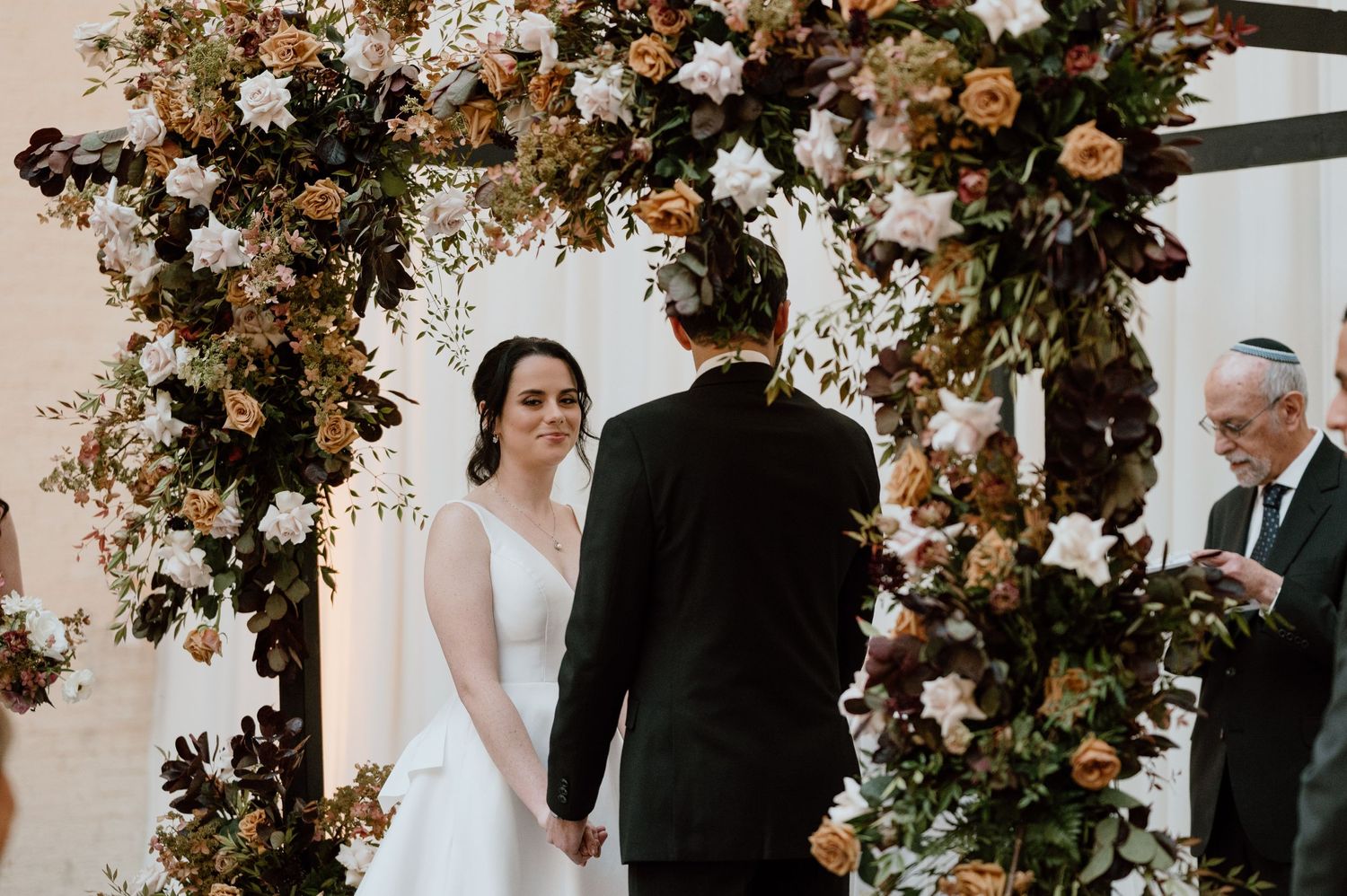 Wedding ceremony under an elegant floral arch with autumn-toned flowers and greenery.