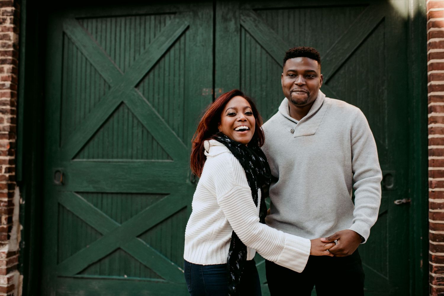 A couple laughs together in front of a green door on a brick building.