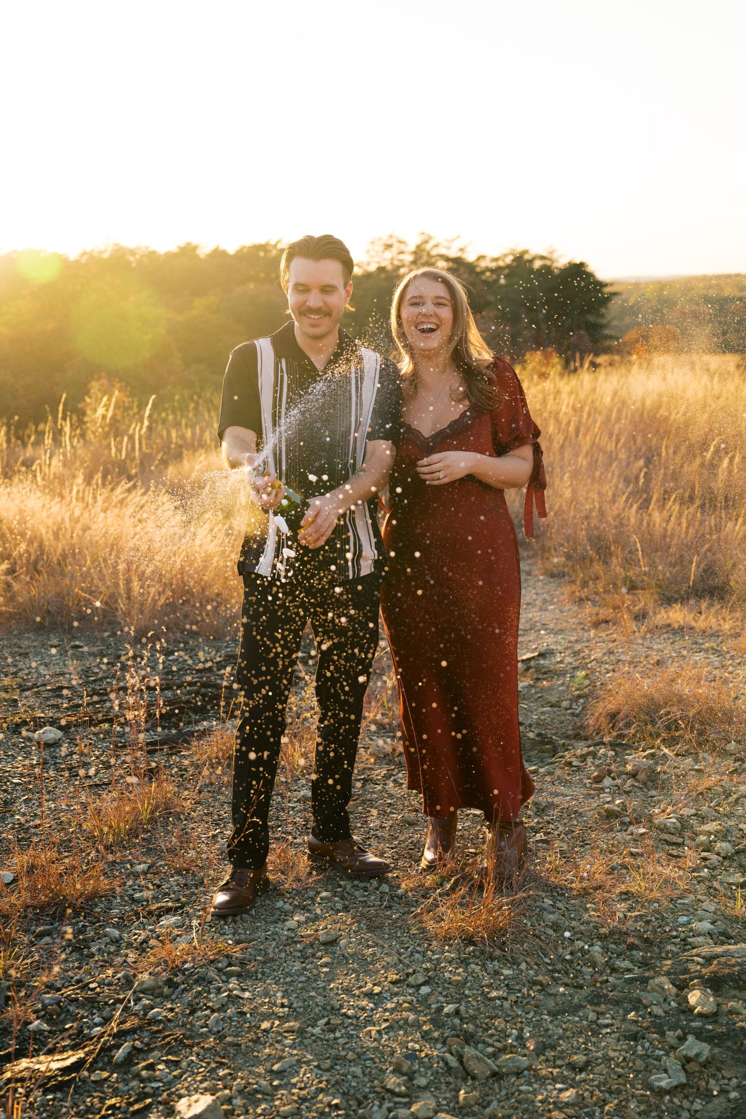 Happy couple laughing and celebrating in a golden field at sunset with champagne spray.