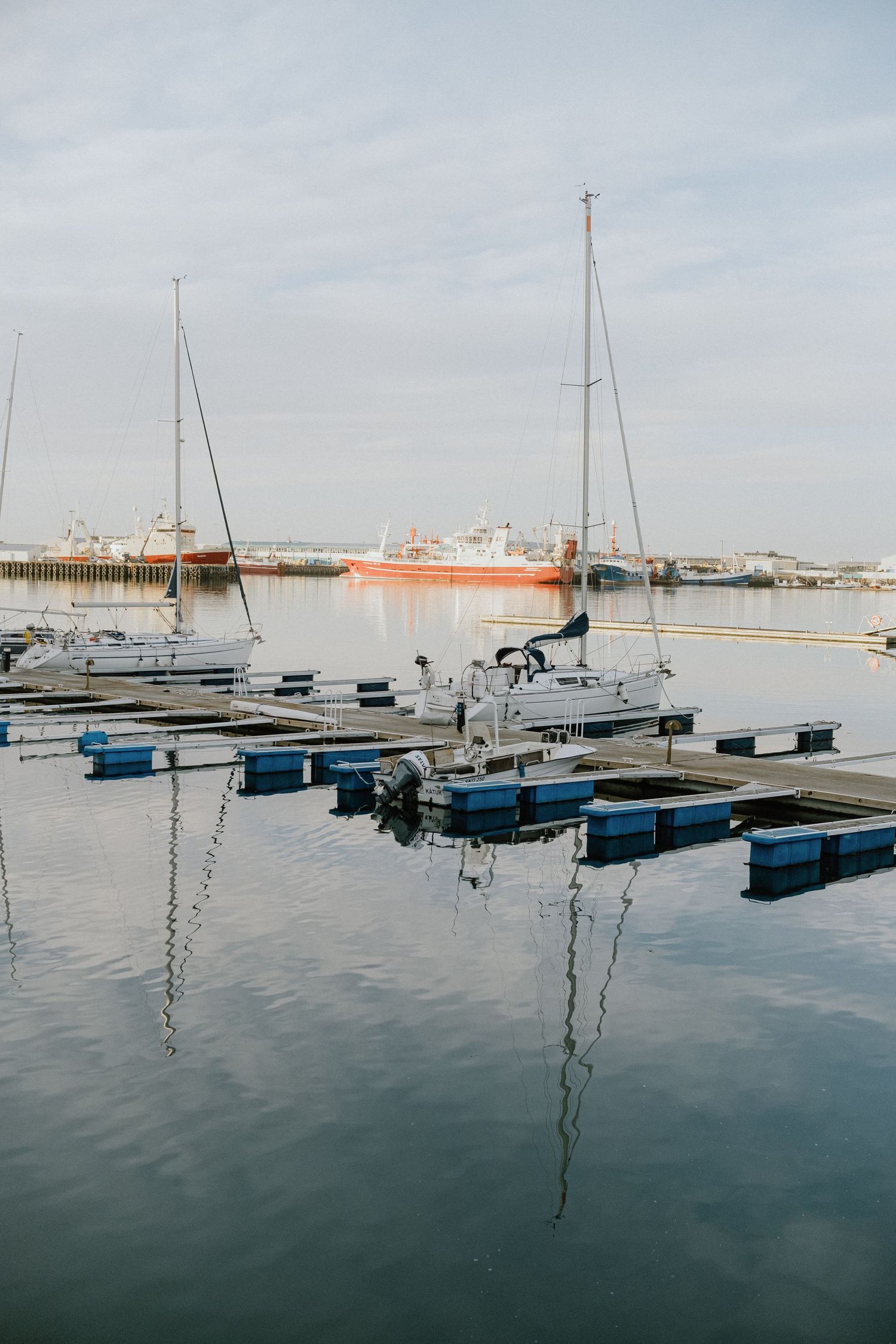 Marina with sailboats and orange fishing vessel reflected in calm harbor waters.