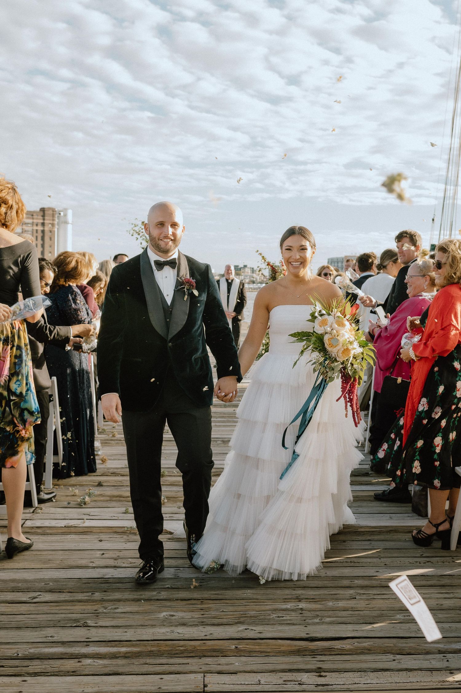 Newlyweds walk down a wooden boardwalk while guests in colorful attire look on.