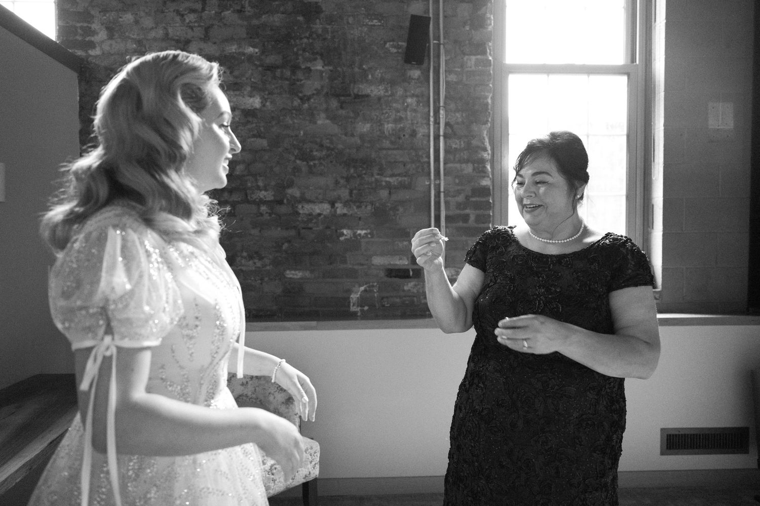 Two people share an animated conversation in a brick-walled room during wedding preparations.