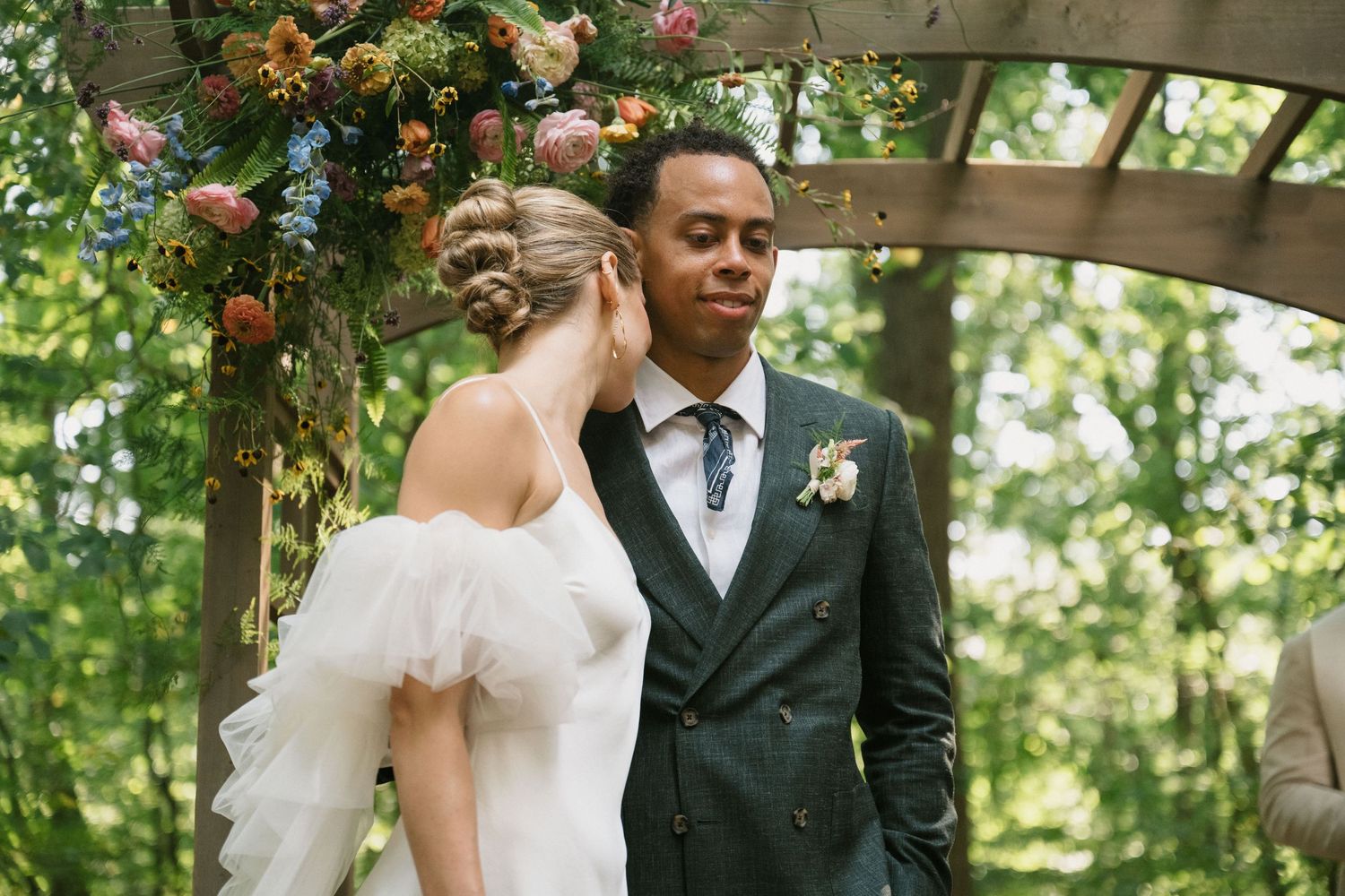 A couple shares an intimate moment beneath a wooden pergola decorated with floral arrangements.