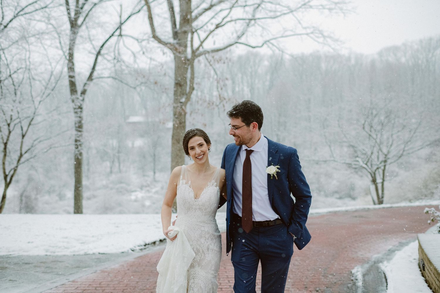 A wedding couple walks together on a snow-covered brick pathway surrounded by winter trees.