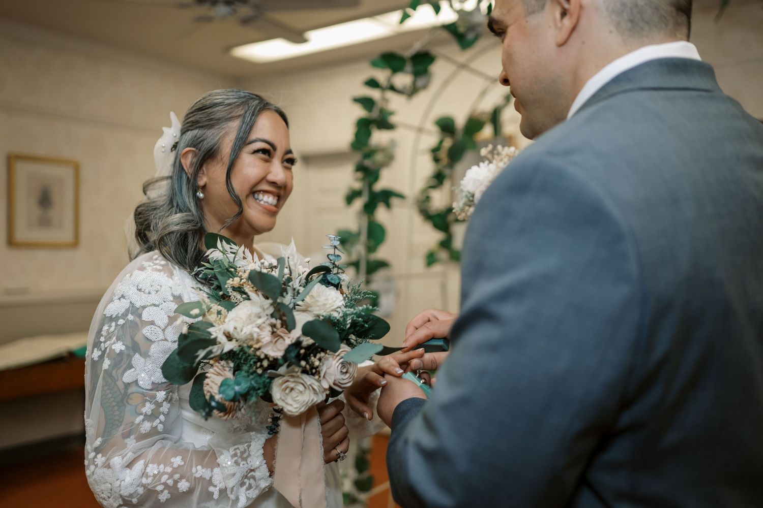 A couple exchanges rings during an intimate indoor wedding ceremony with lace details and a greenery bouquet.
