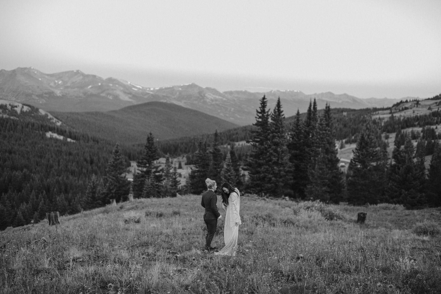 Black and white mountain landscape with two figures standing in a meadow surrounded by evergreen trees.