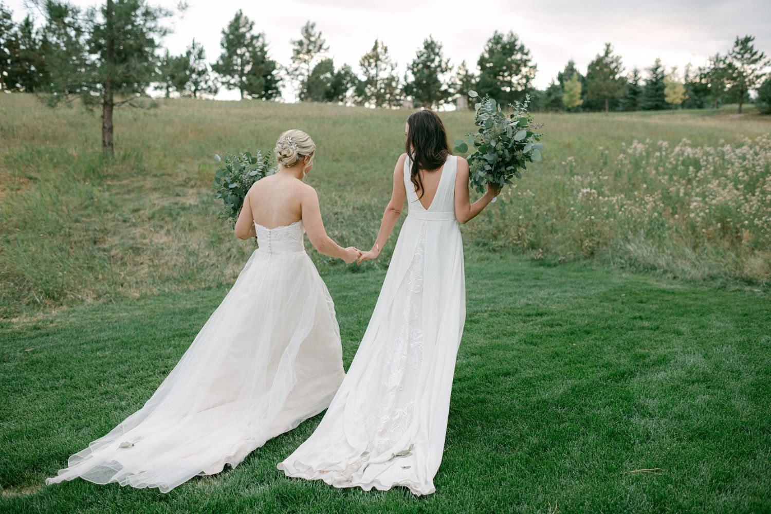 Two people in flowing white wedding dresses hold hands while walking in a grassy field.