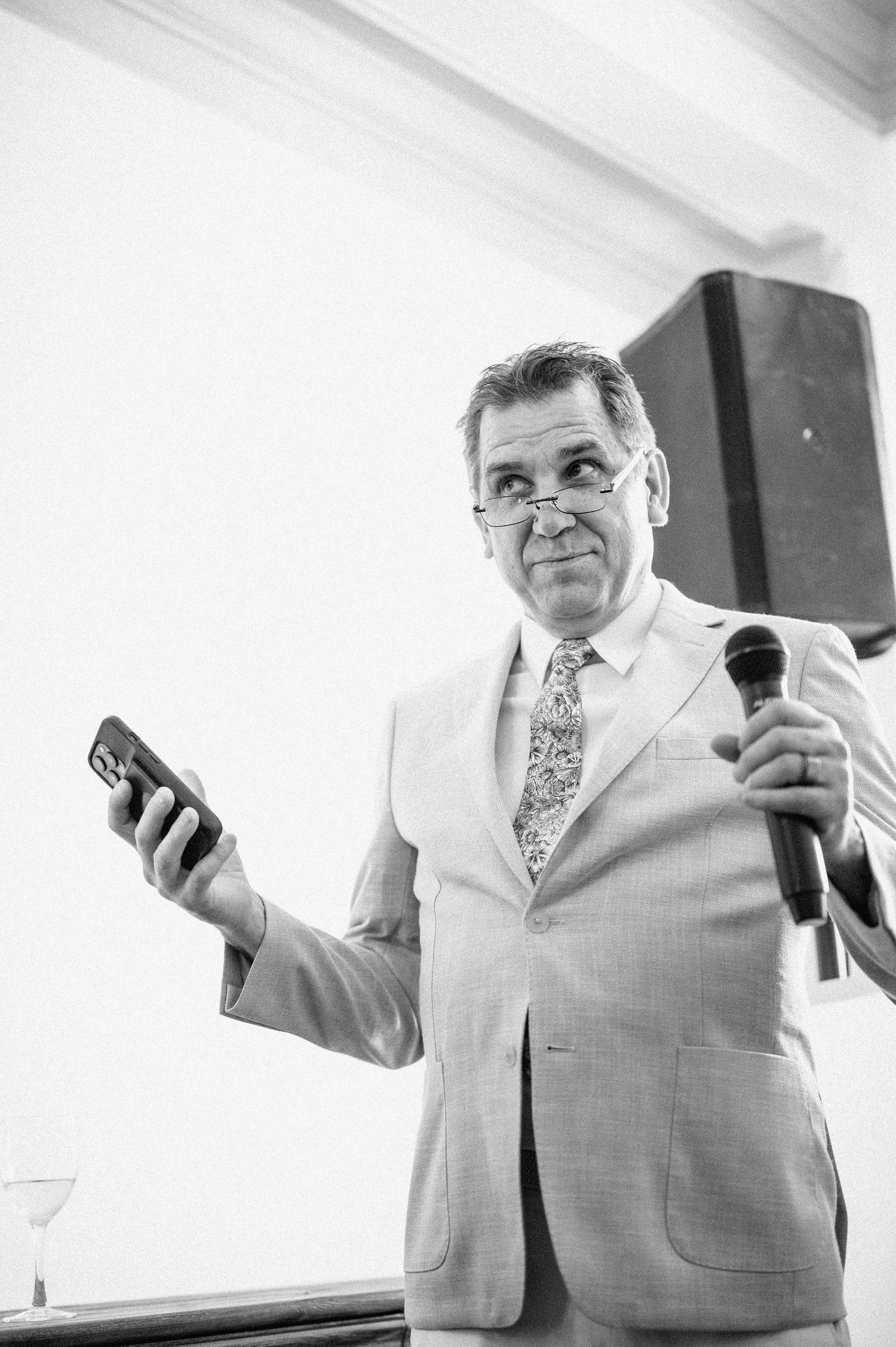Speaker in light colored suit holding phone and microphone during wedding reception.