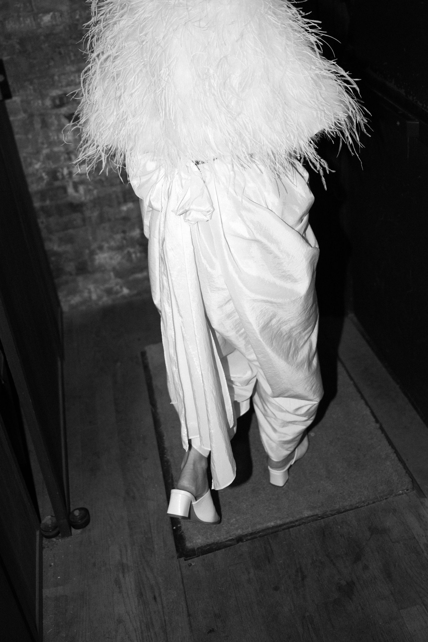 Back view of someone wearing a flowing white dress and fluffy white jacket walking down steps in black and white.