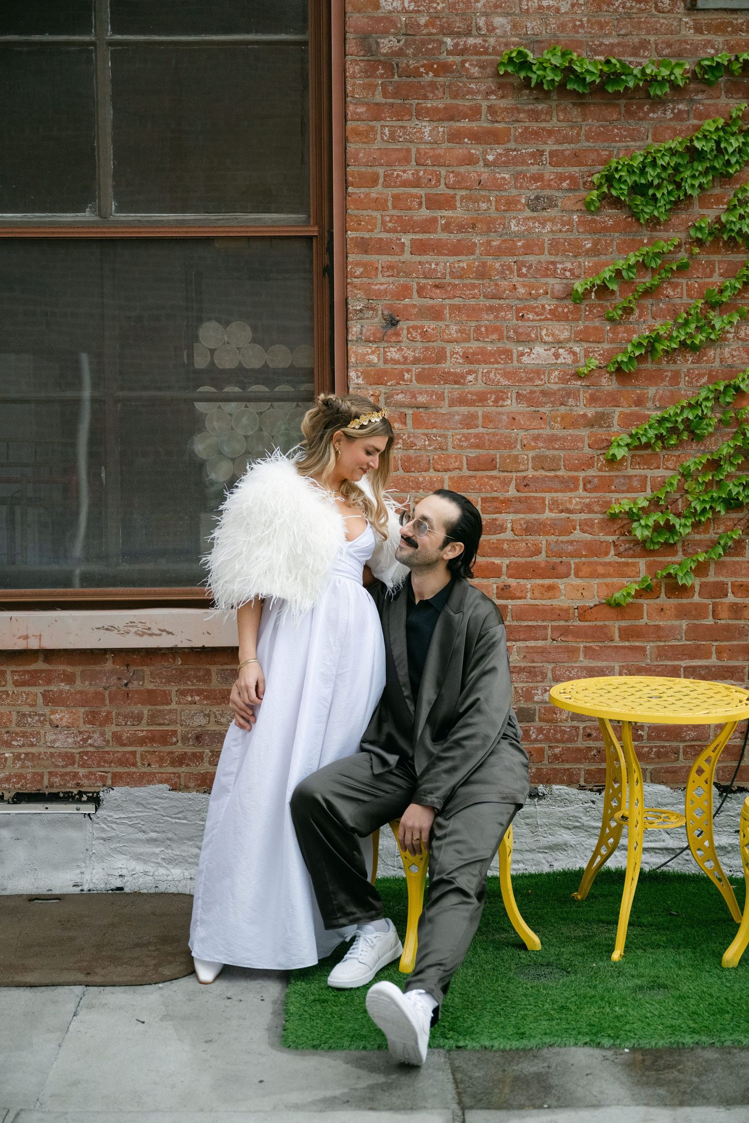 A couple in a courtyard with brick walls, featuring a white dress with feathers and yellow outdoor furniture at Fadensonnen in Baltimore
