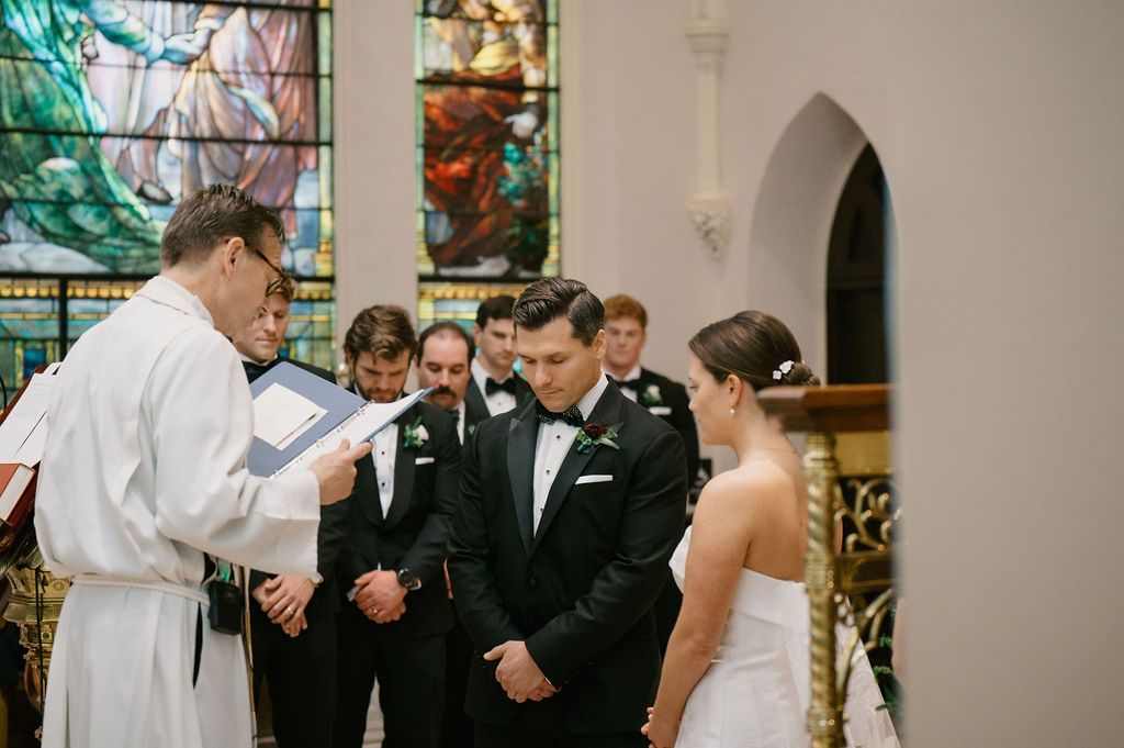 Church wedding ceremony with guests seated in wooden pews beneath arched ceilings and stained glass windows.