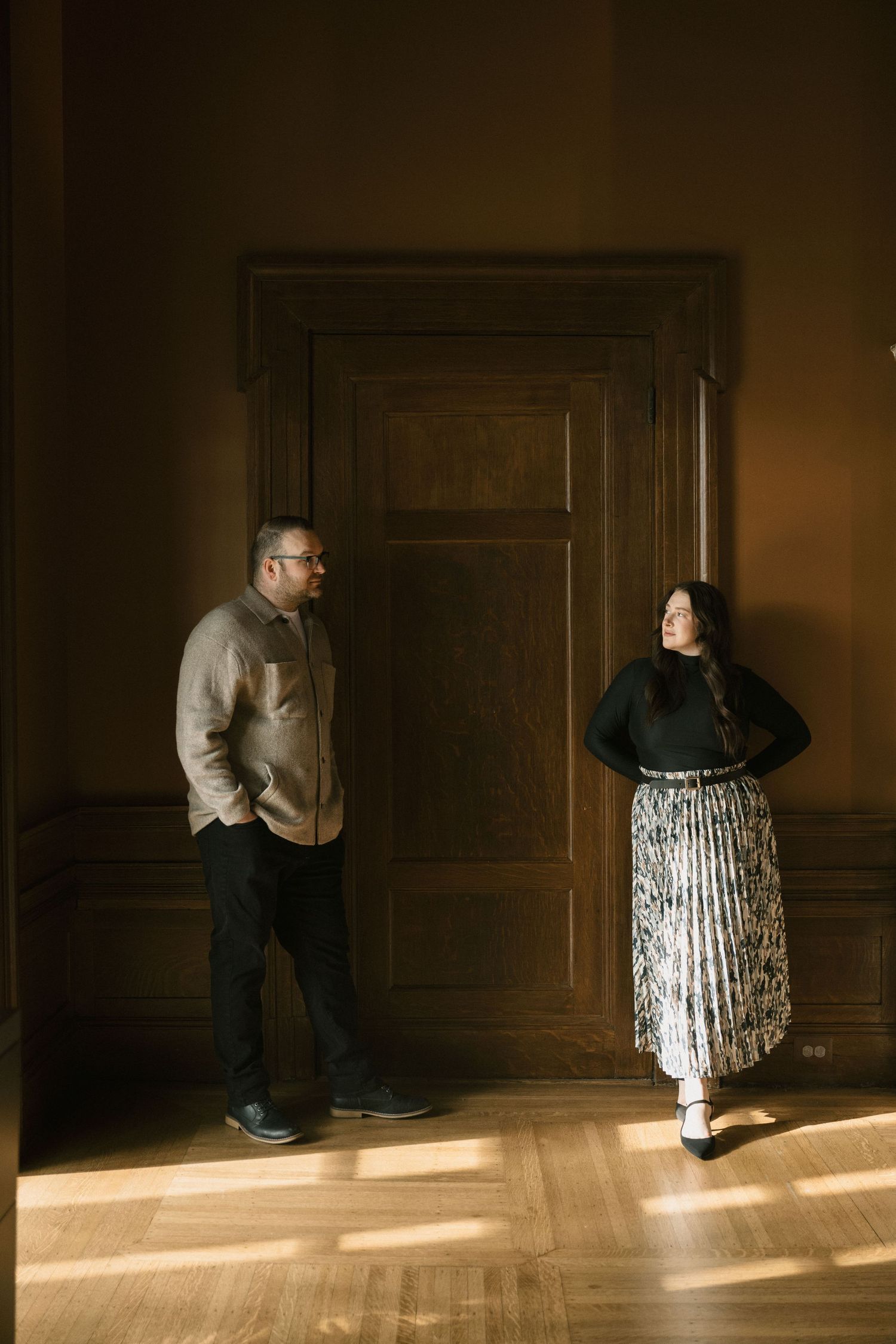 A man and woman stand apart in a dimly lit elegant room with wood paneling and hardwood floors.
