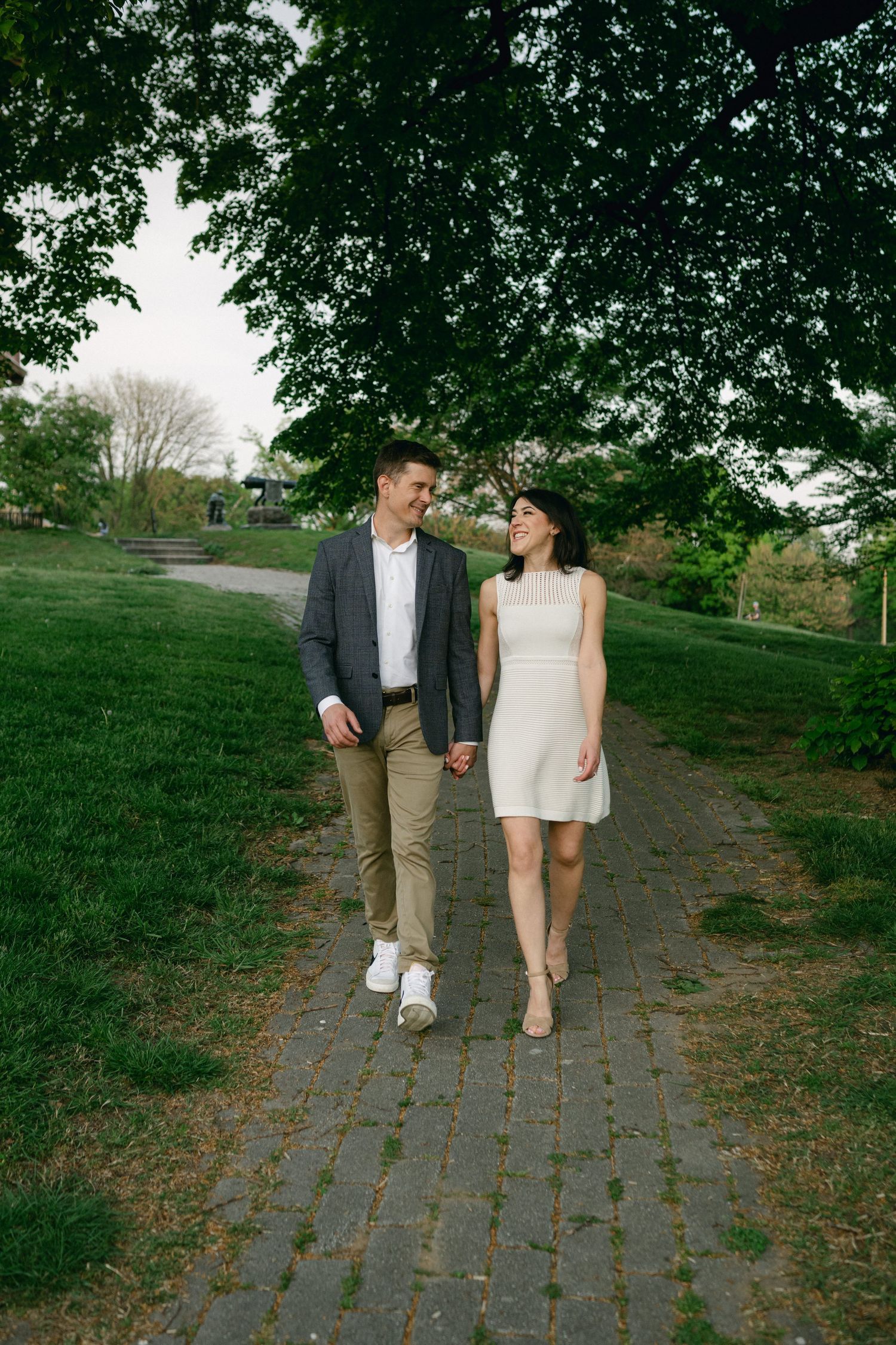 A couple holds hands and walks along a brick path in a lush green park, smiling at each other.