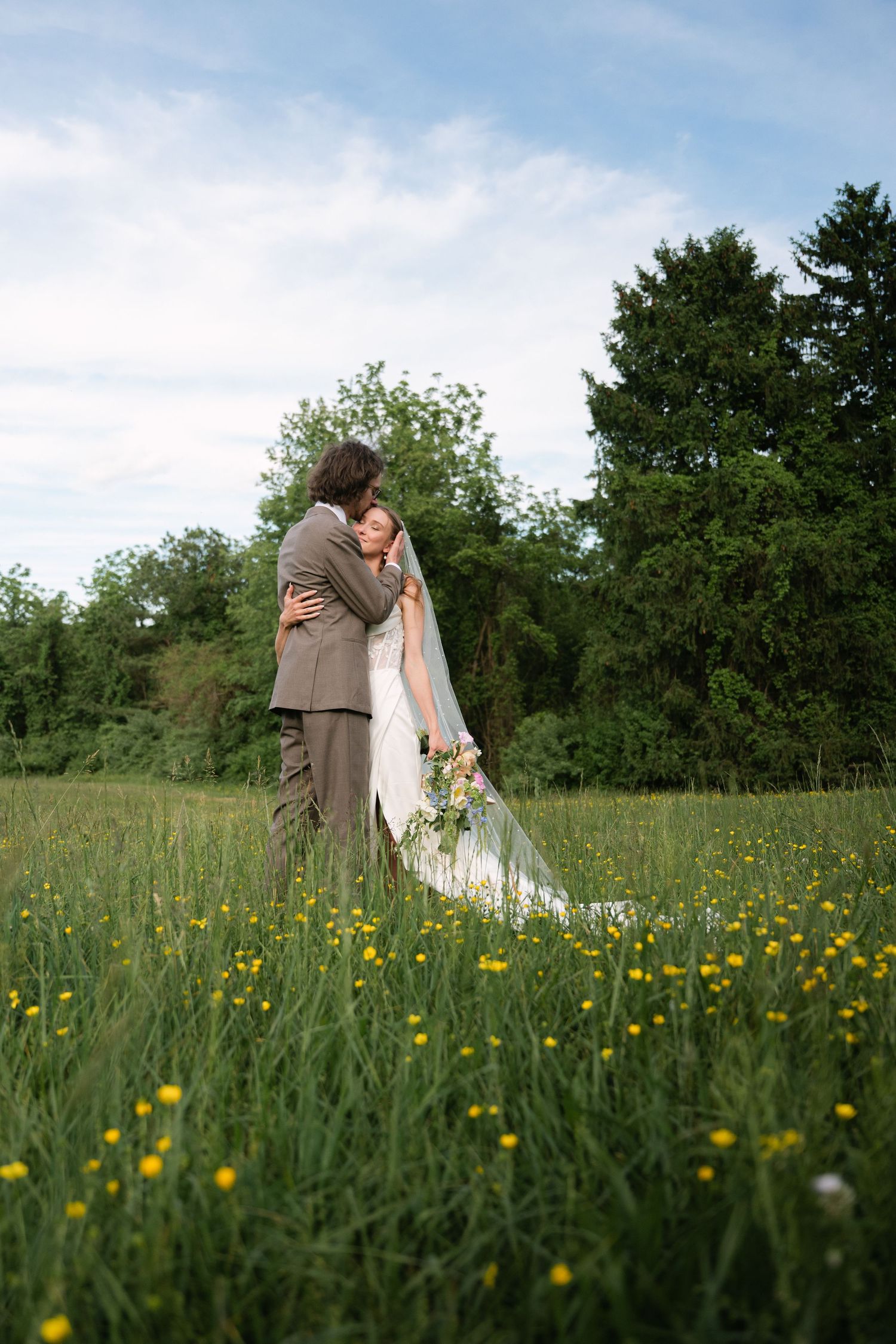 Bride and groom embracing in a lush green meadow with wildflowers, bride wearing a flowing veil and holding a bouquet.