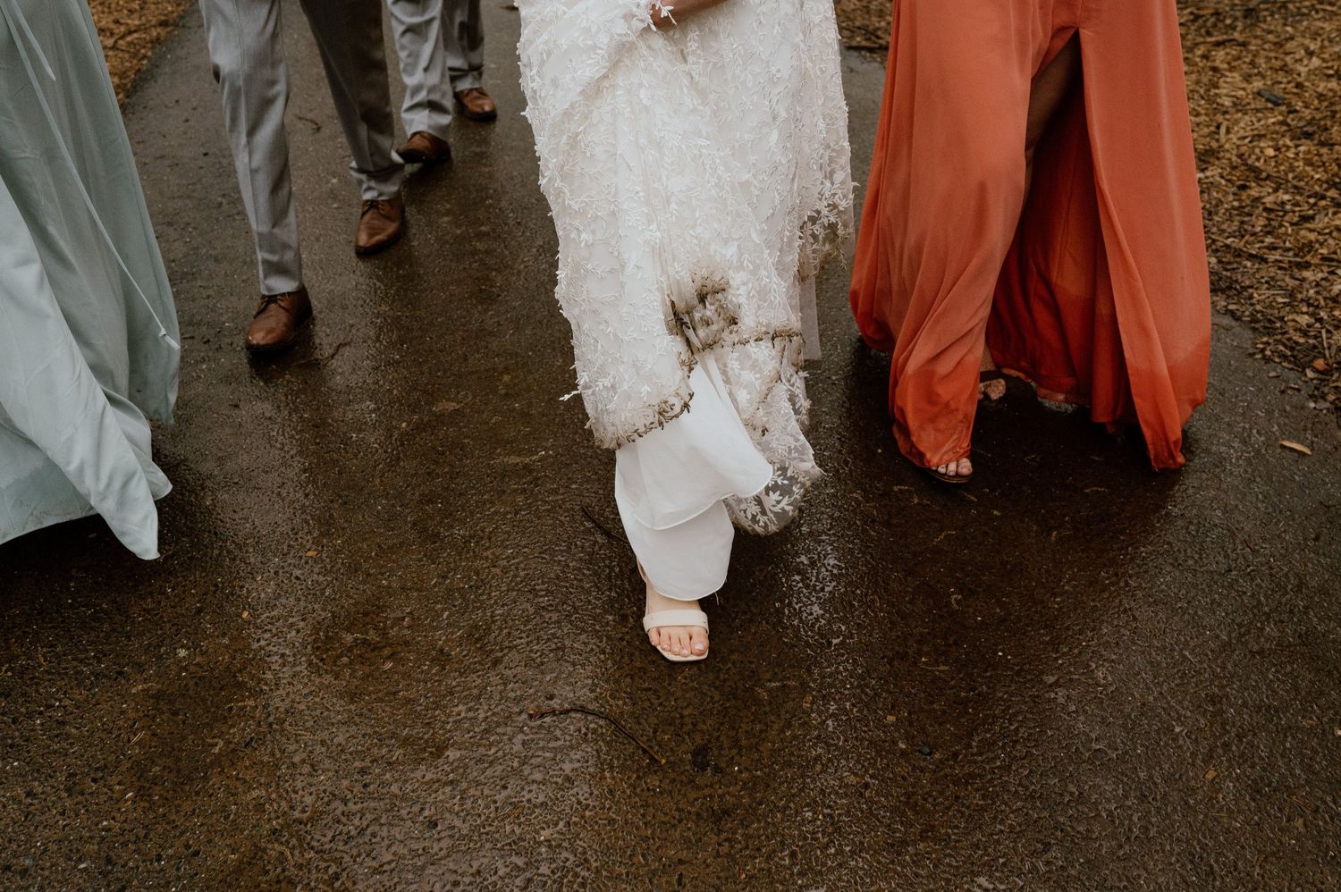 Close-up of wedding attendees walking on wet ground showing dress hem and flowing gowns.